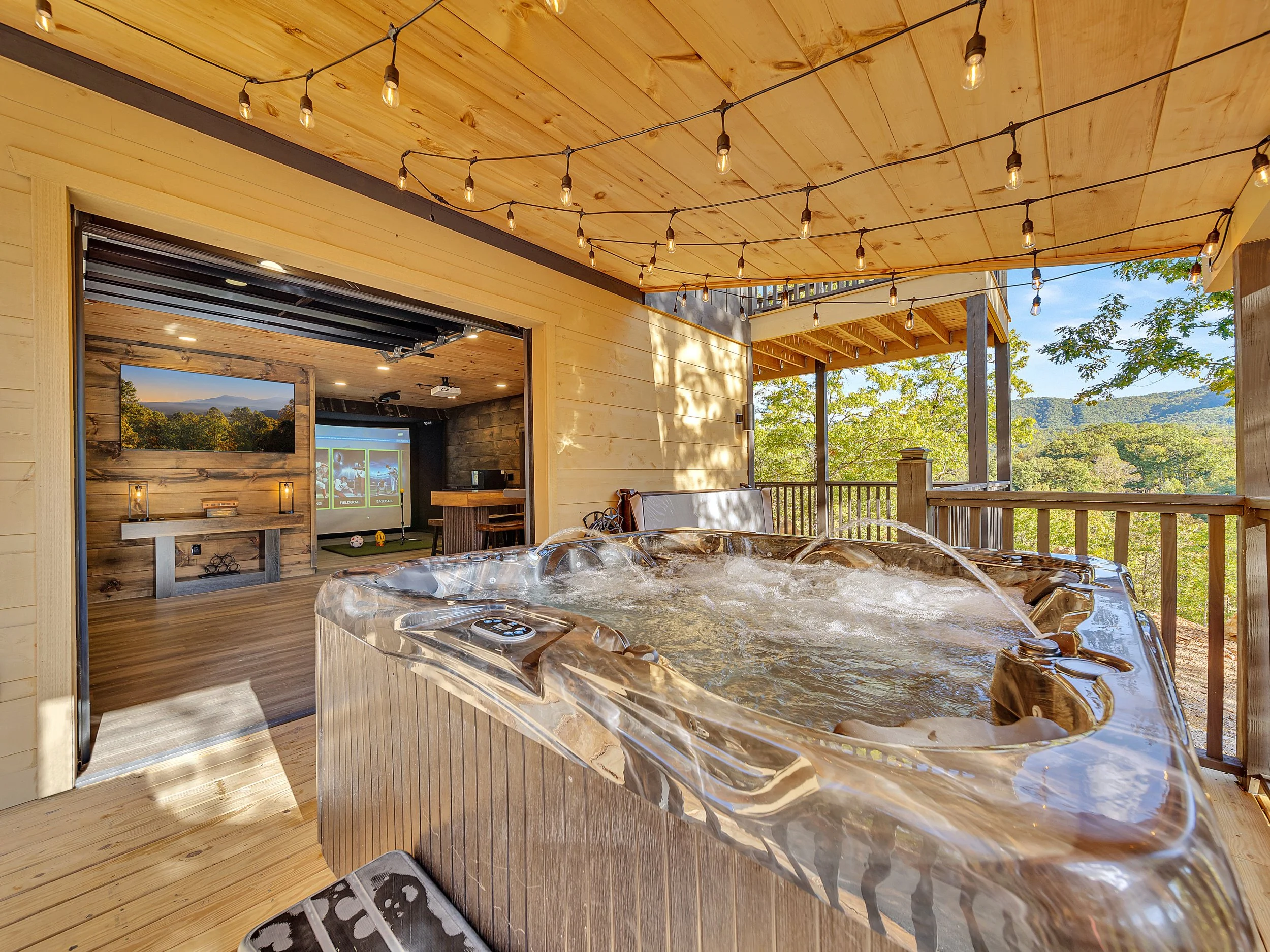 Outdoor deck with a hot tub overlooking a wooded landscape, string lights hanging from the ceiling, and a view of a mountain range in the distance.