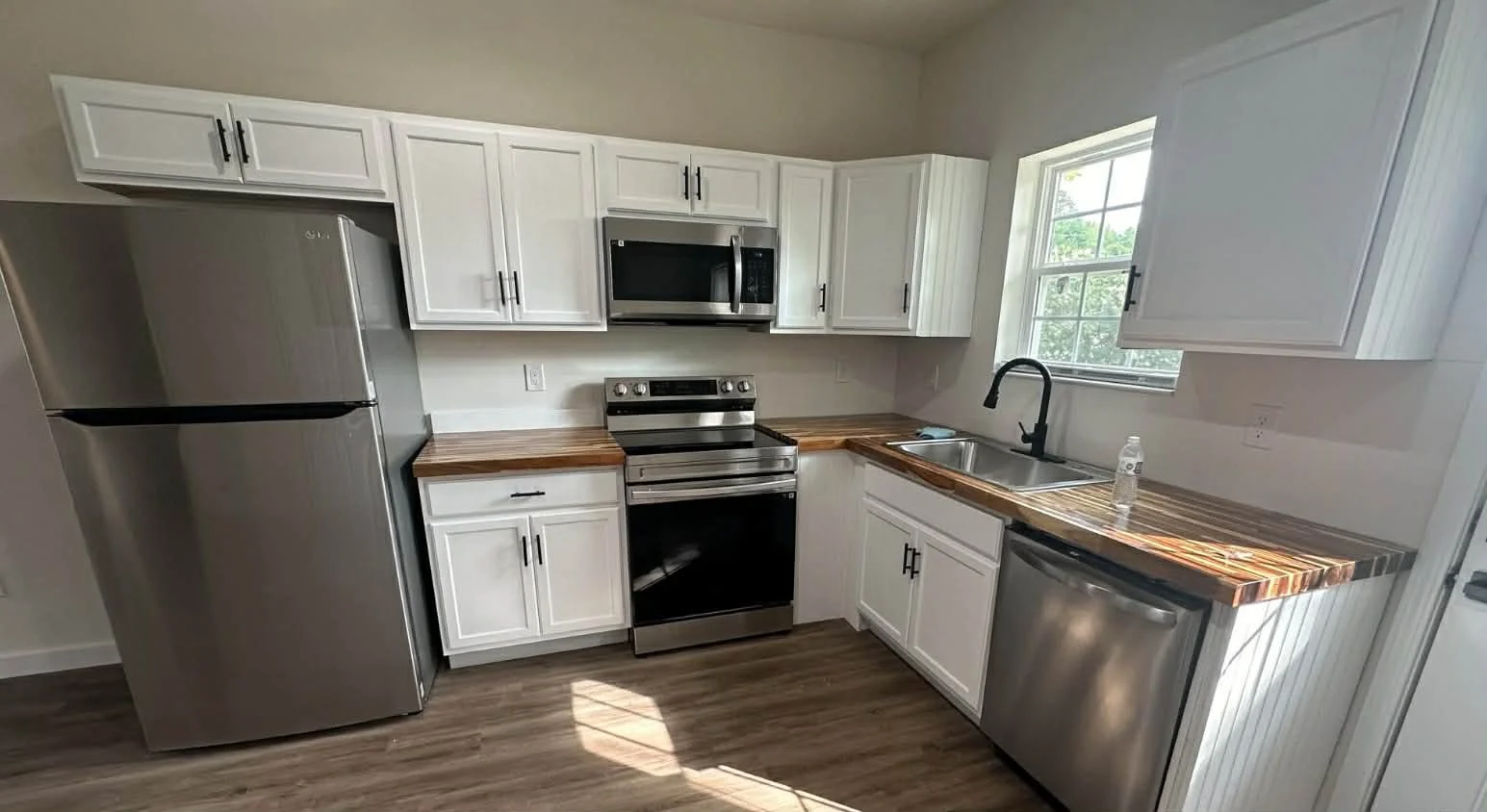 Kitchen with stainless steel refrigerator, microwave, oven, and dishwasher, white cabinets, wooden countertop, and a window above the sink
