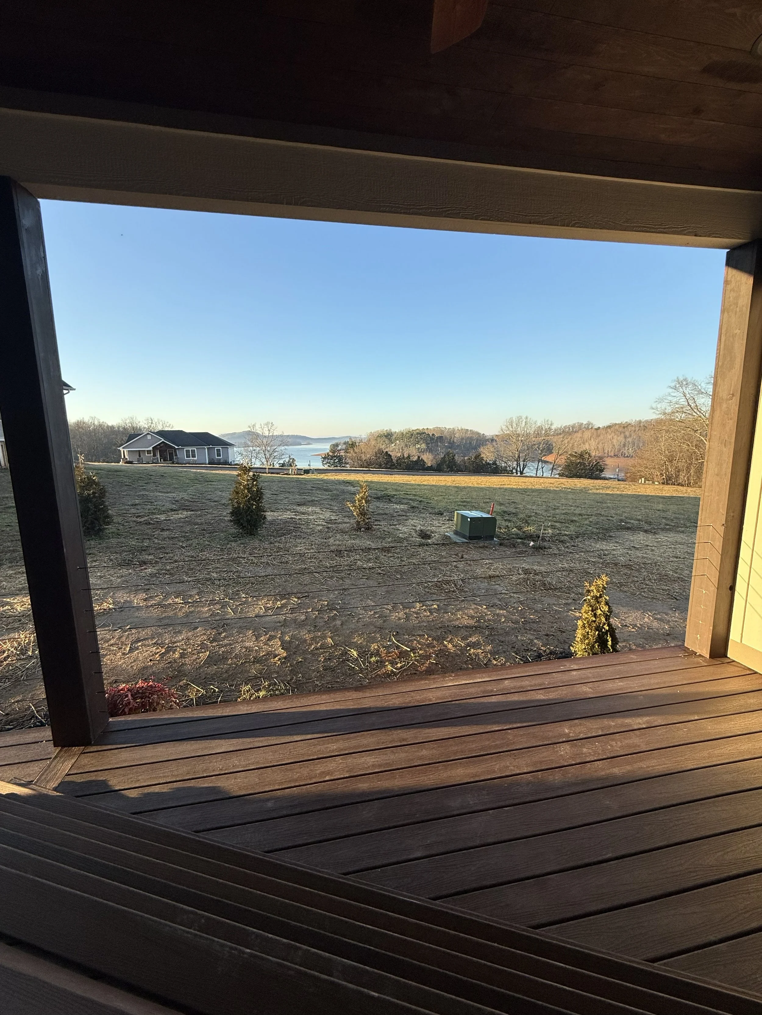 View from a wooden porch looking out over a grassy yard with small bushes, a house in the distance near a body of water, and a clear blue sky.