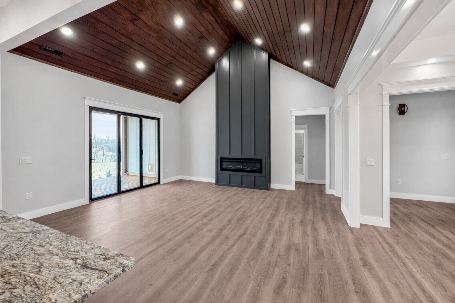 Empty living room with hardwood floors, white walls, a dark wood vaulted ceiling with built-in lighting, a modern black fireplace, and sliding glass doors leading to a balcony.