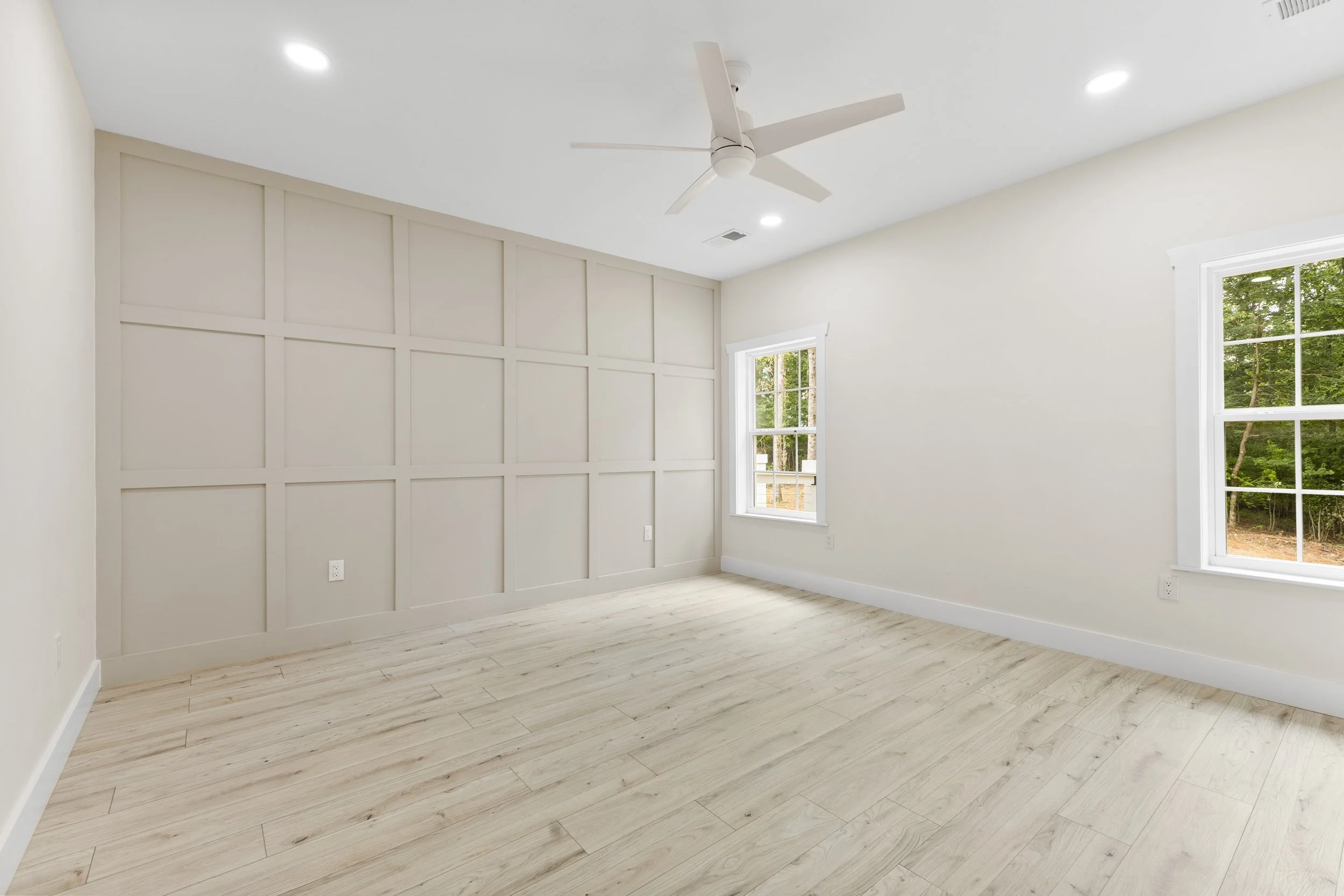 Empty bedroom with light wood flooring, off-white walls, two windows, white ceiling fan, and recessed lighting.