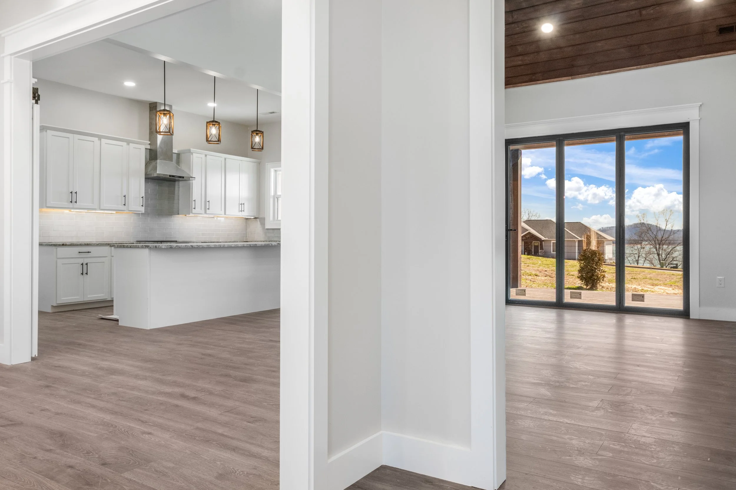 Empty modern kitchen and living space with large floor-to-ceiling window showing an outdoor scene with houses, trees, and mountains.