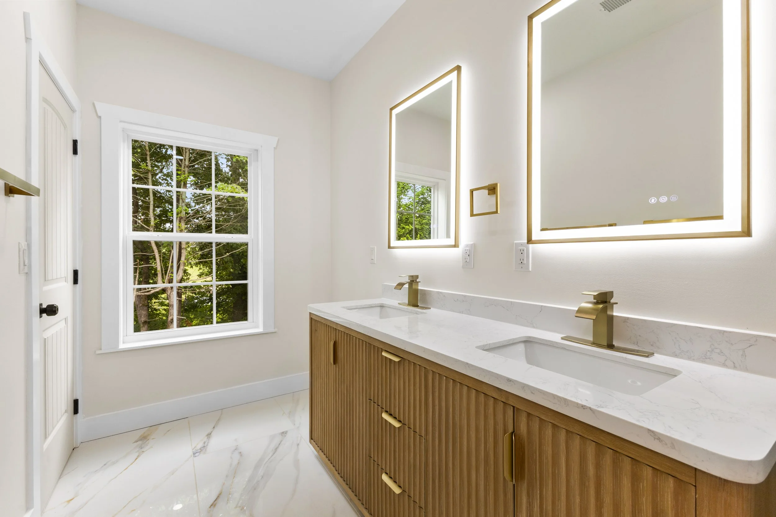 Modern bathroom with dual sinks, wooden cabinet, white marble countertop, and large mirrors with LED lighting, next to a window showing green trees outside.