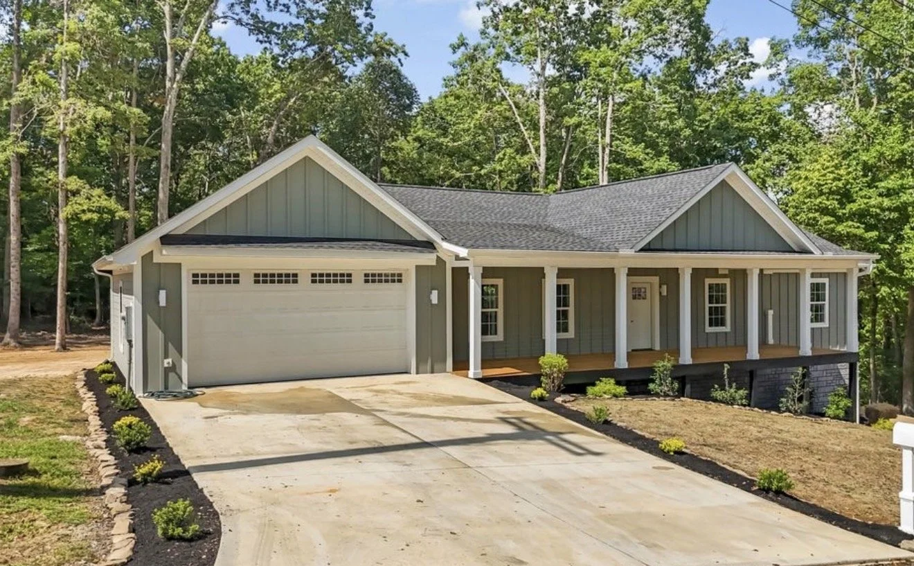 A newly built, light gray house with a front porch, attached two-car garage, and a landscaped yard surrounded by trees.