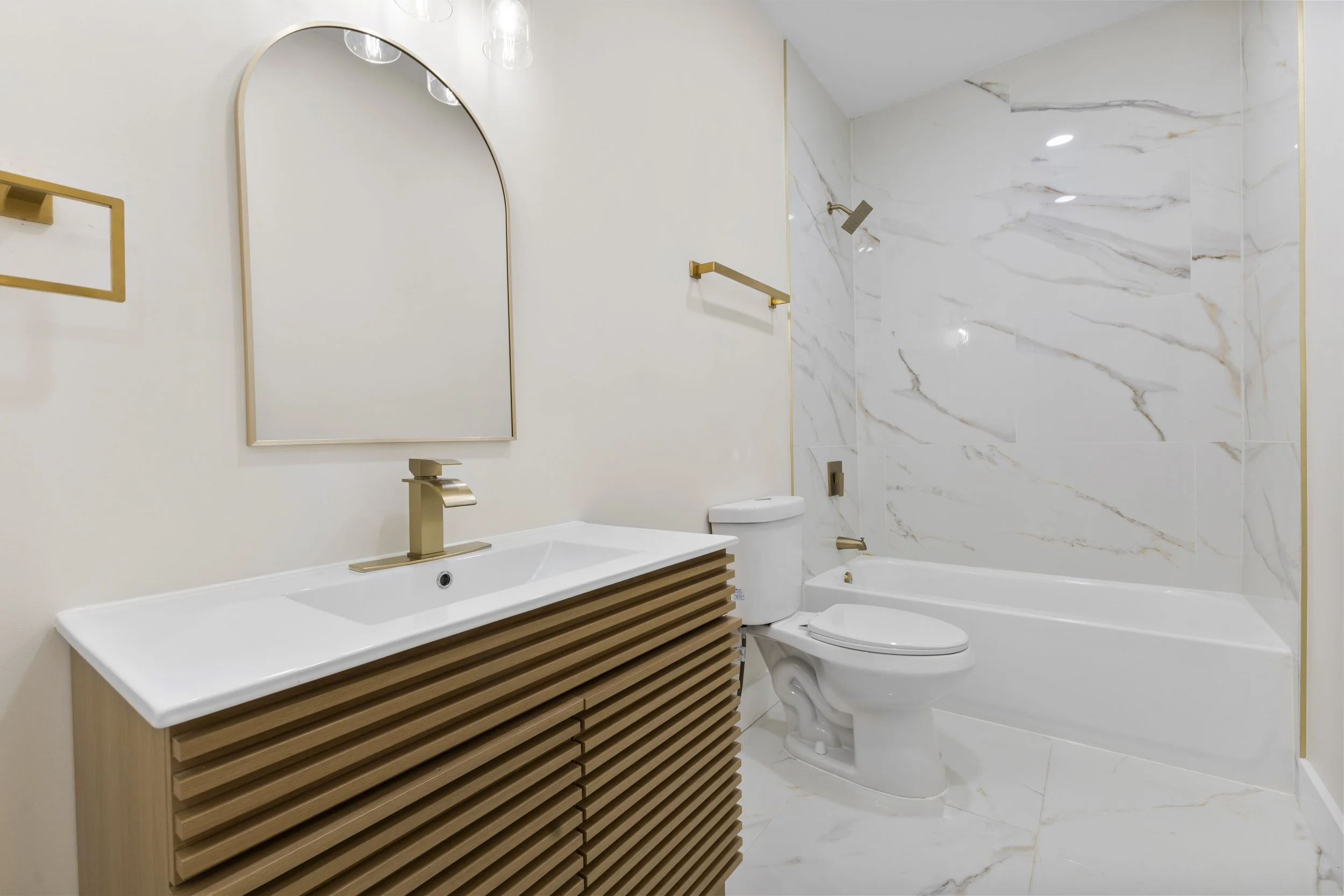 Modern bathroom with a wooden vanity, white sink, brass fixtures, a mirror, a toilet, a bathtub, and marble tiles.