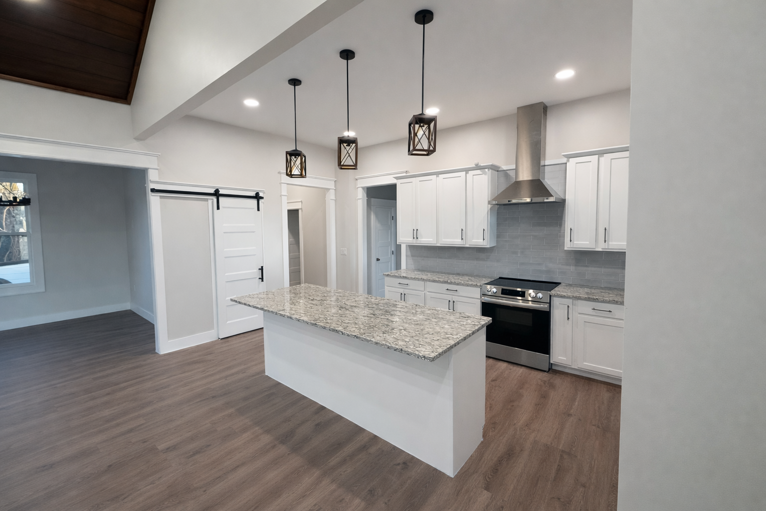 Modern kitchen with white cabinets, granite countertops, island, stainless steel oven, range hood, gray tile backsplash, wooden floors, hanging pendant lights, white walls, and a barn door.