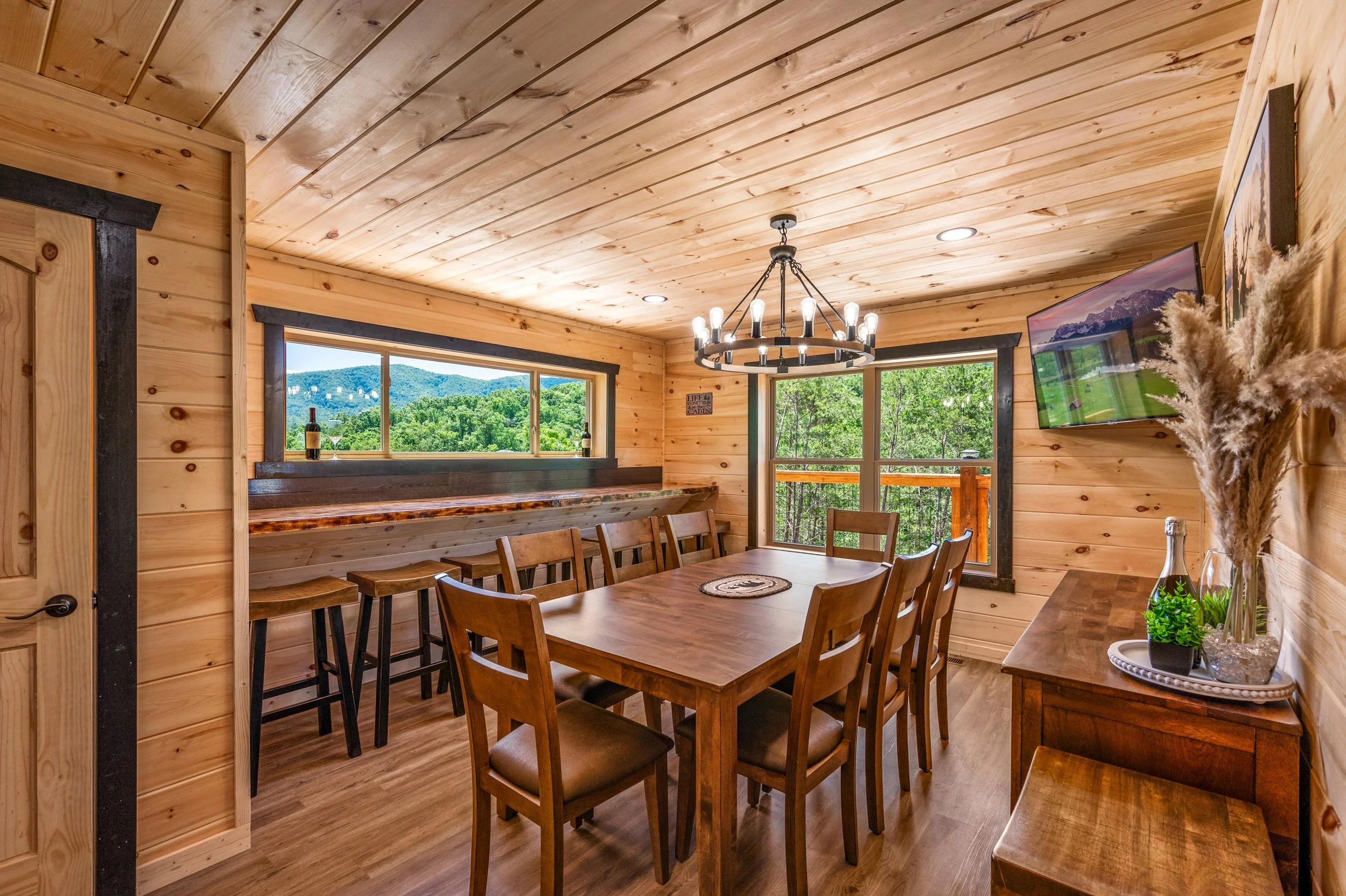 Interior view of a wooden dining room with a large dining table, eight chairs, a chandelier, a sideboard with decorative plants, and windows showing a green outdoor landscape.
