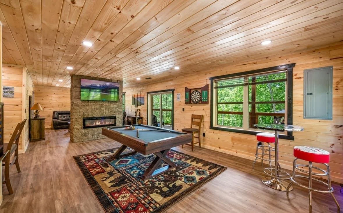 Living room with wood-paneled walls and ceiling, featuring a flat-screen TV above a stone fireplace, a pool table on a patterned rug, and seating area with a leather chair and side table. Large windows reveal green trees outside, and a small bar area