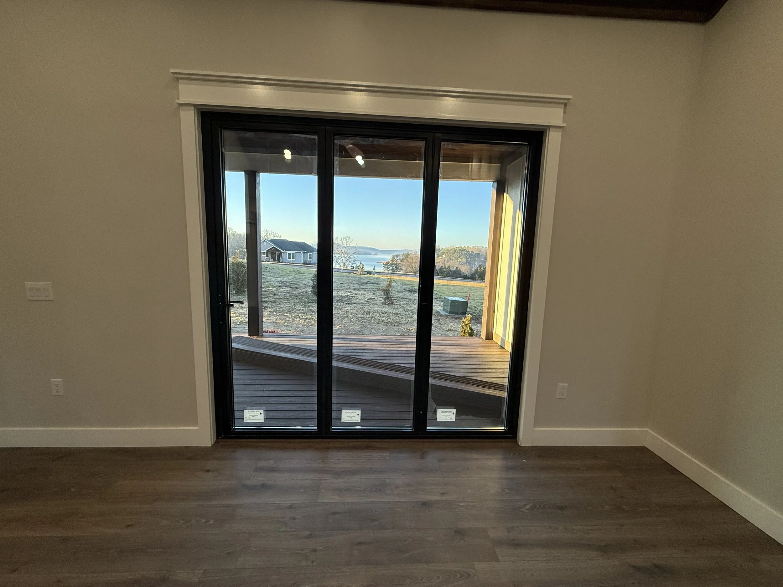 Empty room with sliding glass door leading to a wooden deck, overlooking a yard with a house and lake in the background.