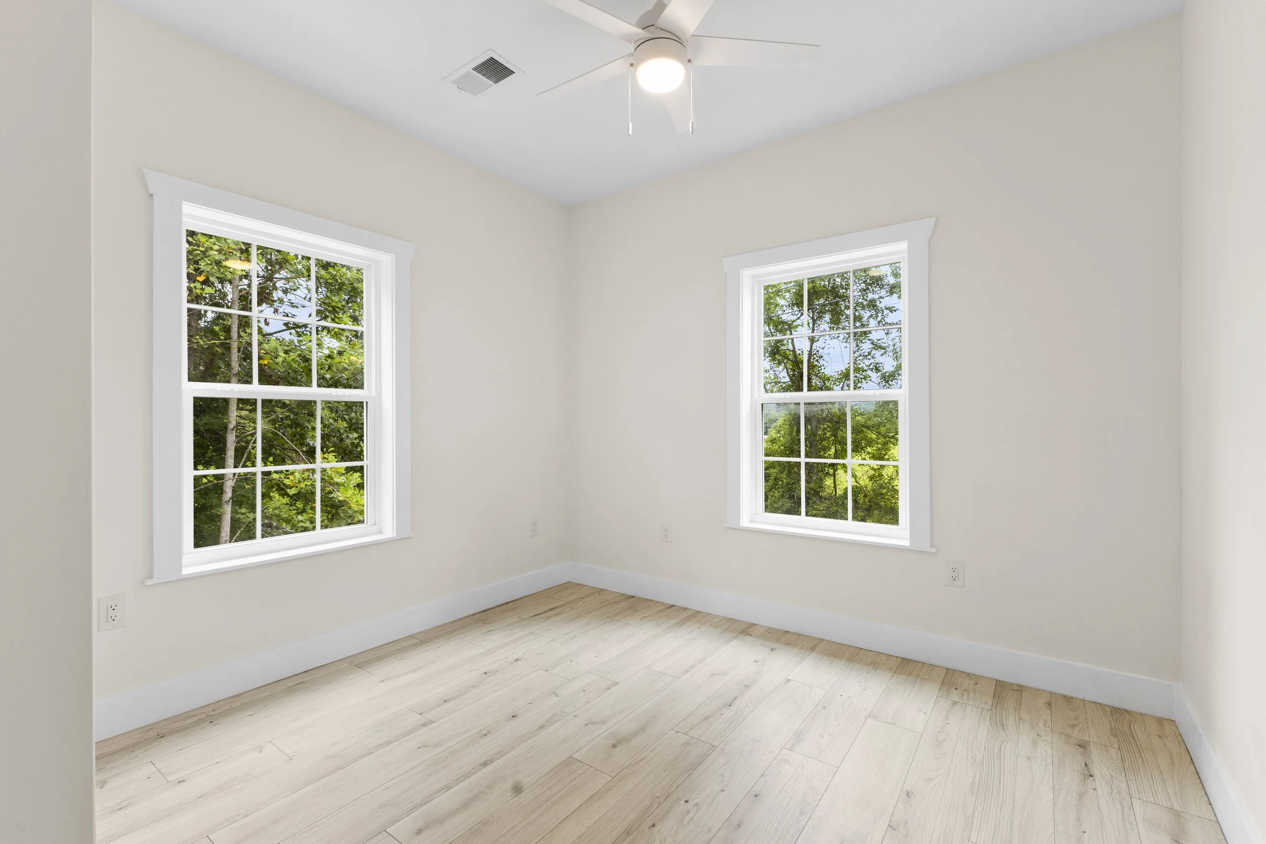 Empty room with two large windows showing green trees outside, light-colored hardwood floors, and white walls with baseboards.