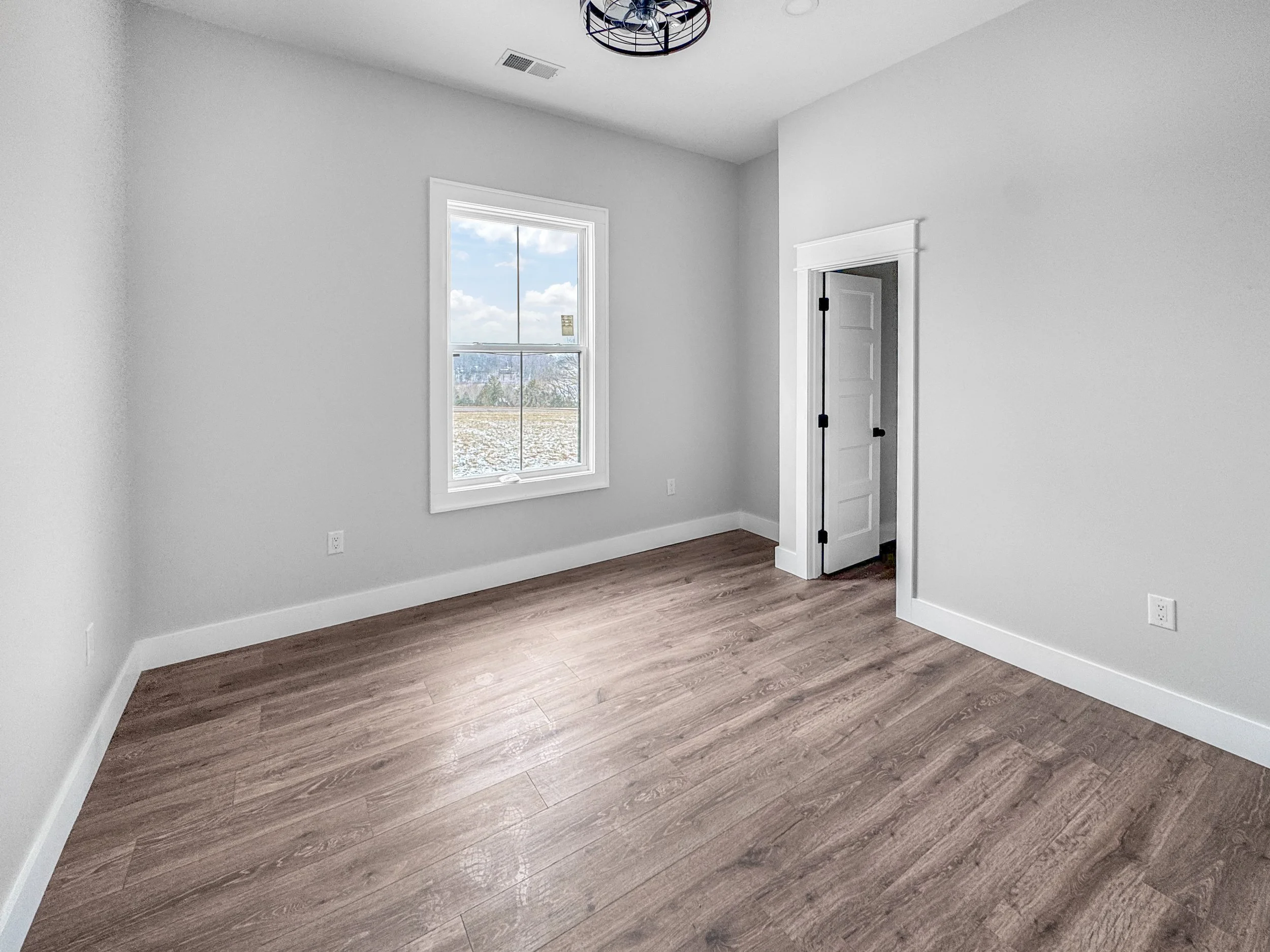 Empty room with white walls, wooden flooring, a window showing a scenic view outside, and an open door leading to another space.