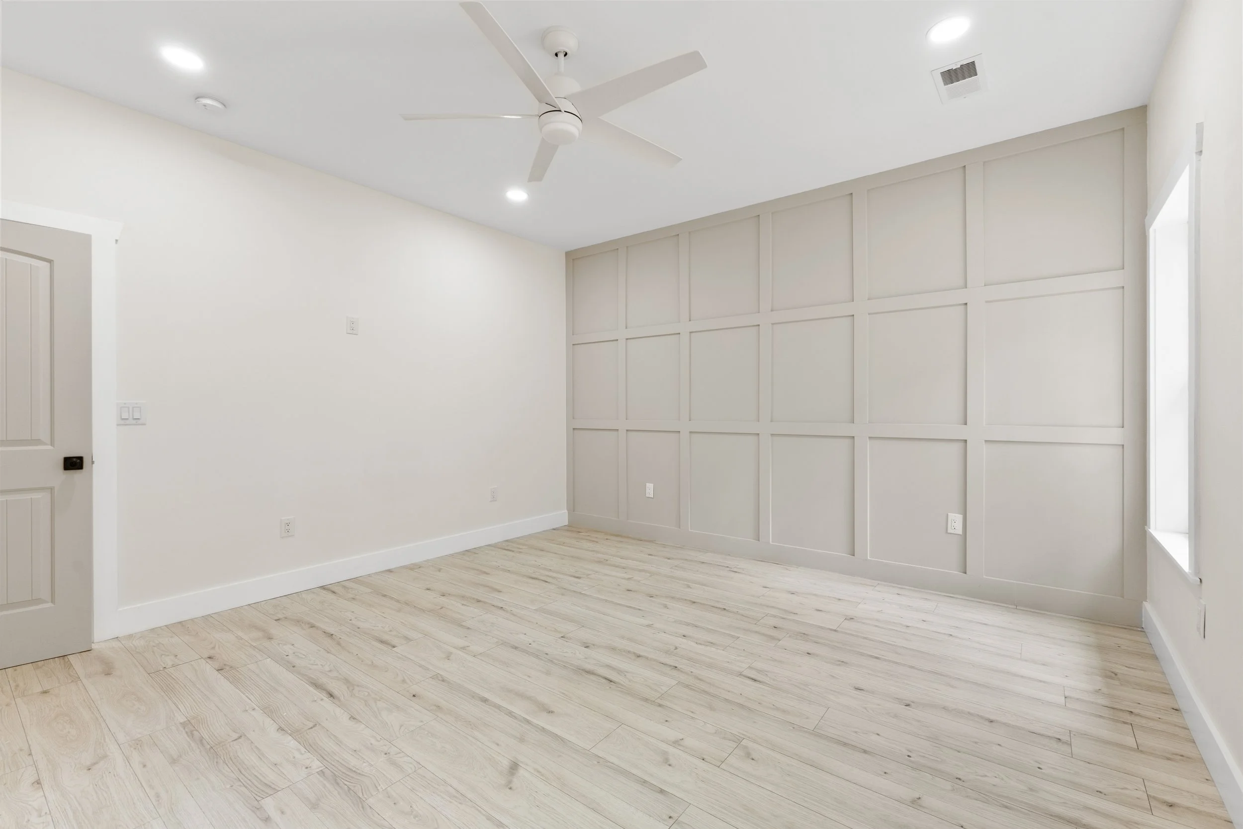 Empty room with white walls, wood flooring, a ceiling fan, and a large window, with one wall featuring decorative paneling.