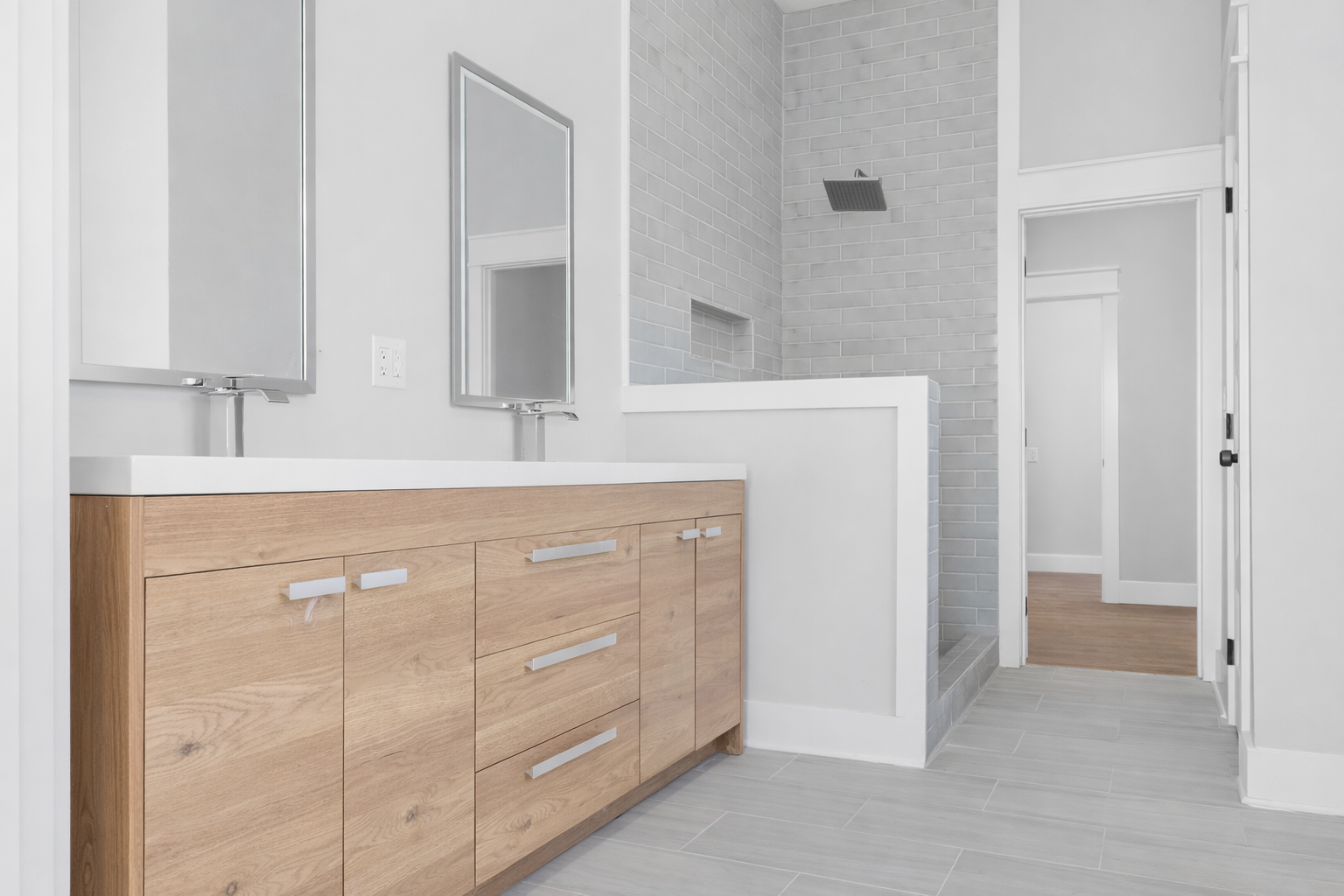 Modern bathroom with a wooden vanity, two mirrors, gray tiled shower with a rain showerhead, and a doorway leading to a room with wooden flooring.