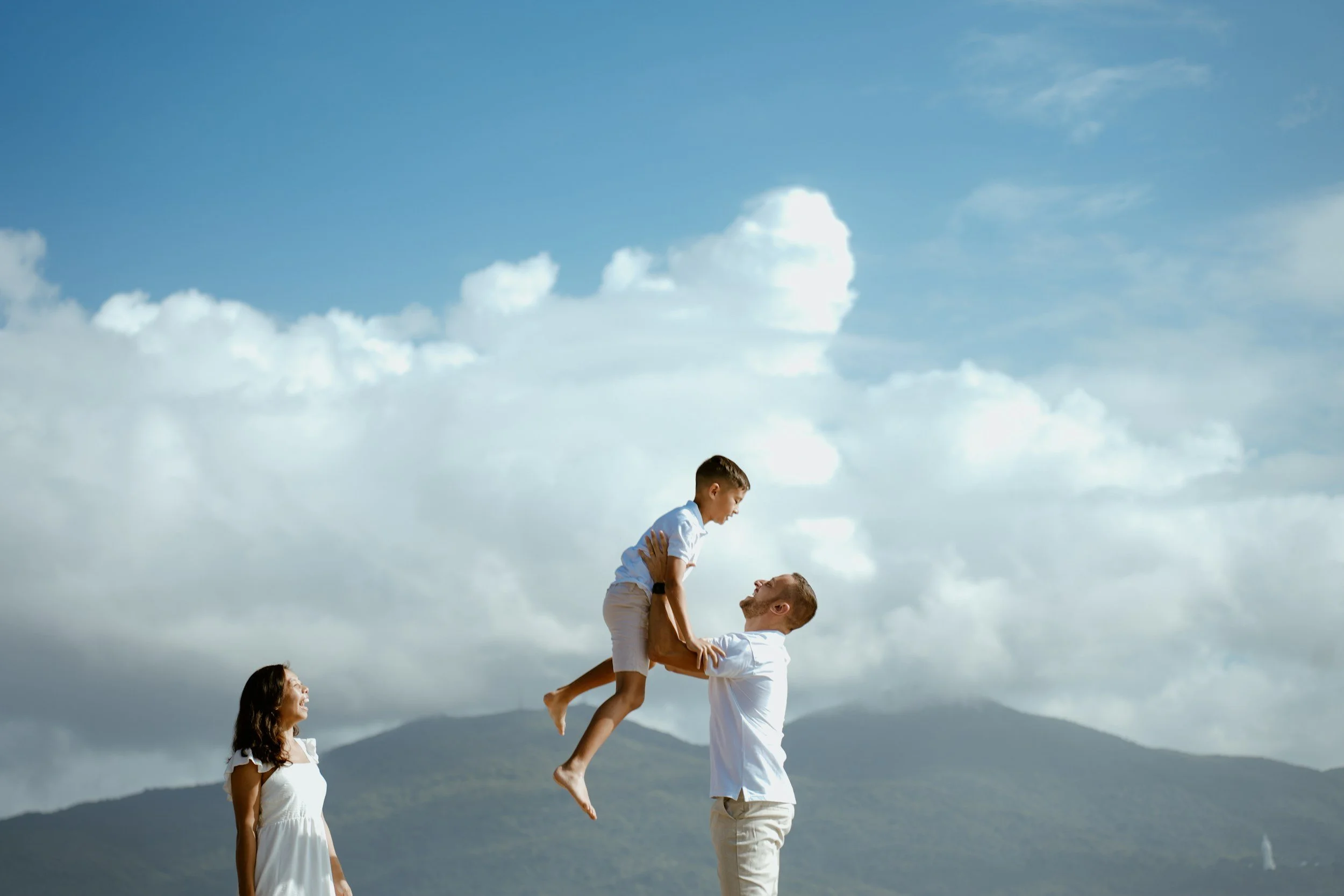 A man lifts a boy into the air while a woman stands nearby, outdoors with mountains and cloudy sky in the background.