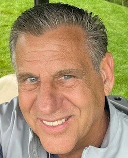 Close-up of a smiling middle-aged man with short gray hair, blue eyes, and a light complexion, outdoors with green grass in the background.