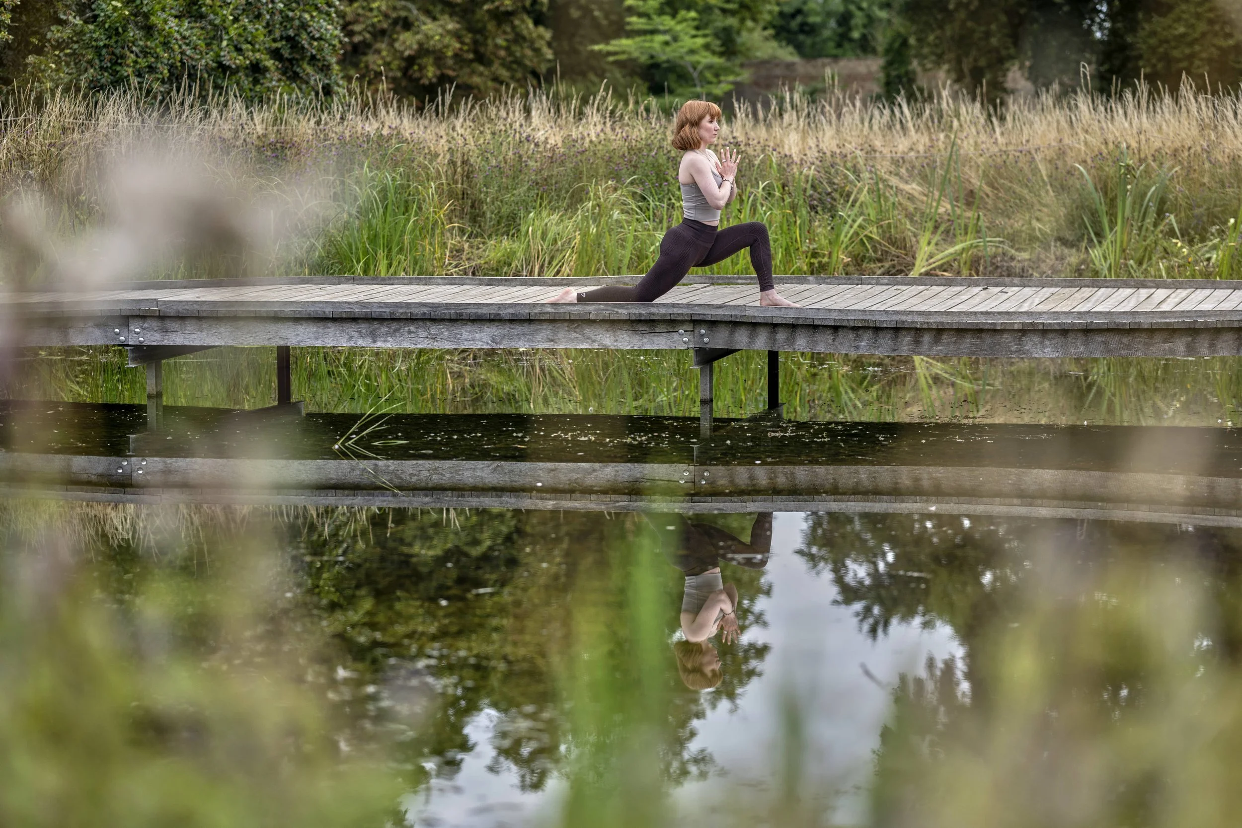 Saffron Walden Yoga Teacher, in mid yoga flow holding a low lung pose with hands in prayer mudra at heart centre. On a bridge near Saffron Walden and reflected in the pond below.