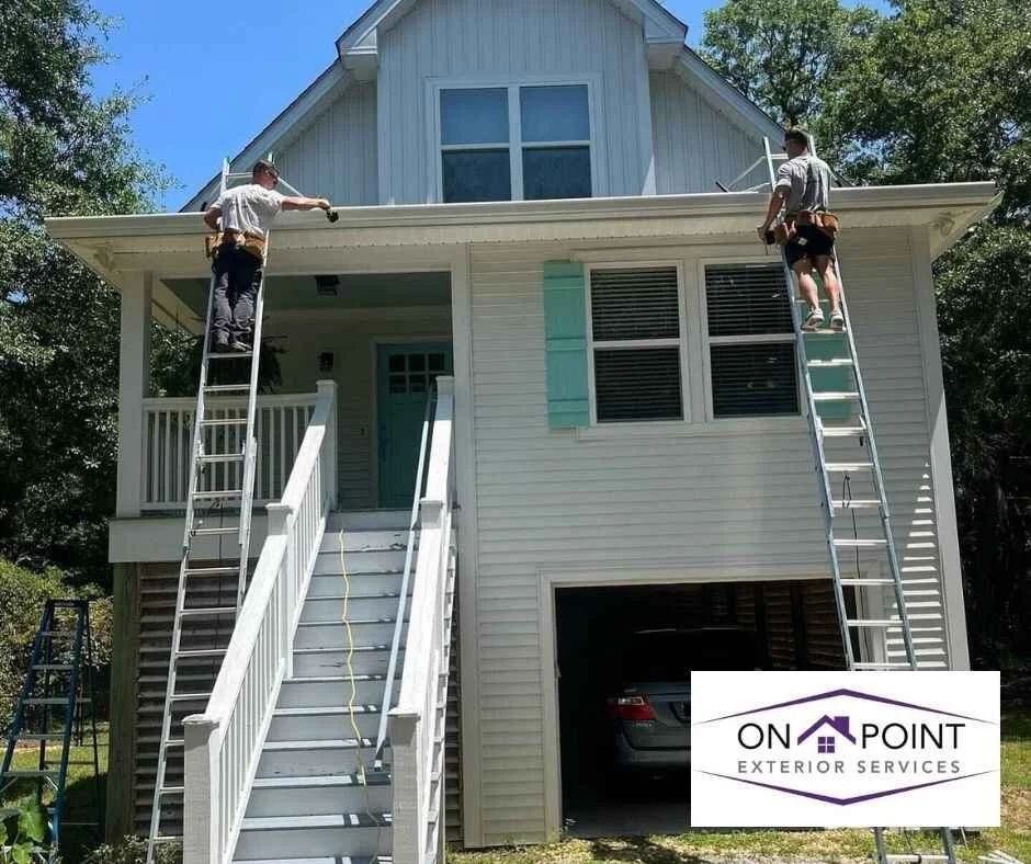 Two workers are on ladders installing or repairing the roof of a two-story house with white siding. A garage is visible at the bottom. There is a construction company sign that reads 'On Point Exterior Services' in the front yard.