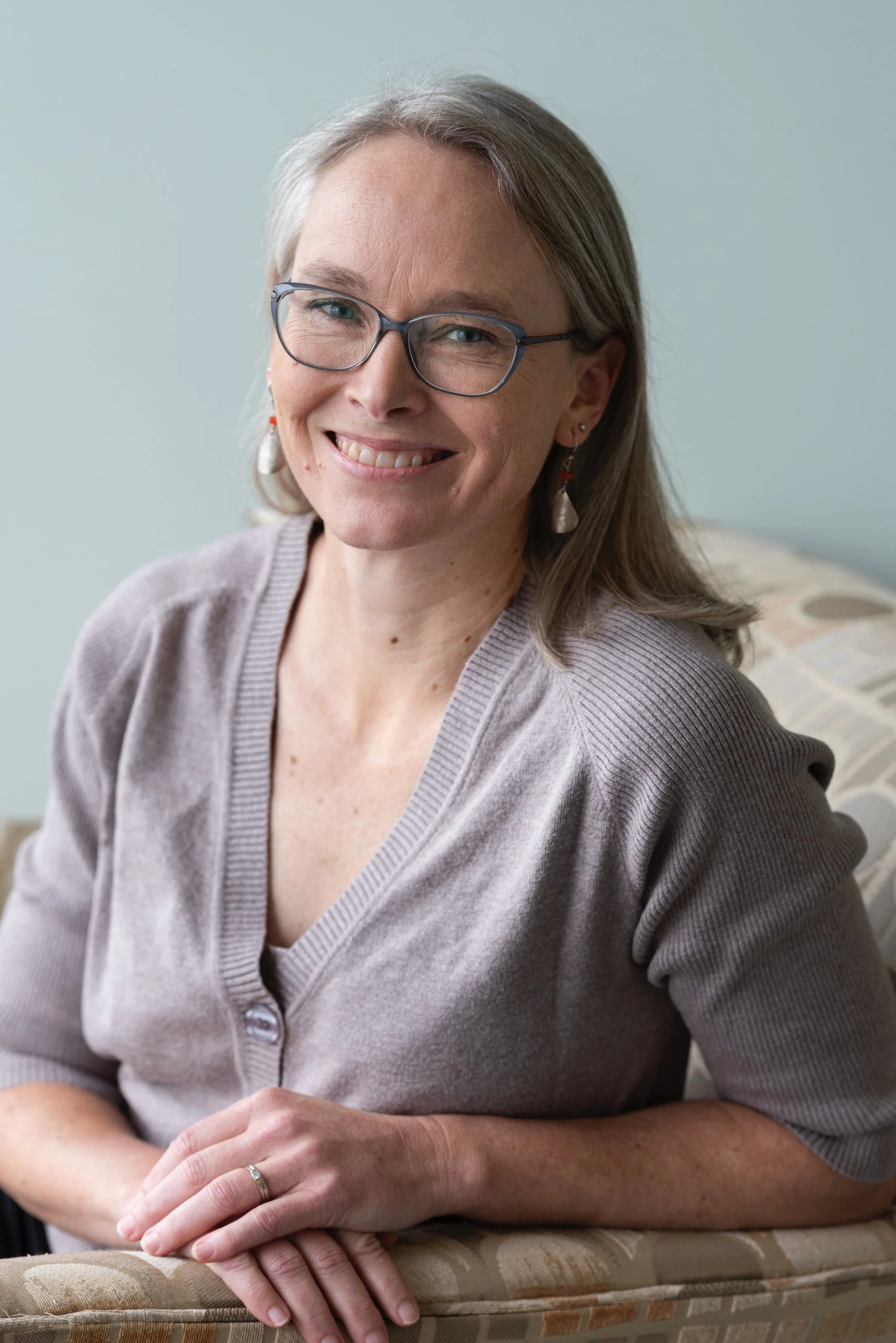 A woman with glasses and earrings, smiling, sitting on a patterned chair with her hands clasped.