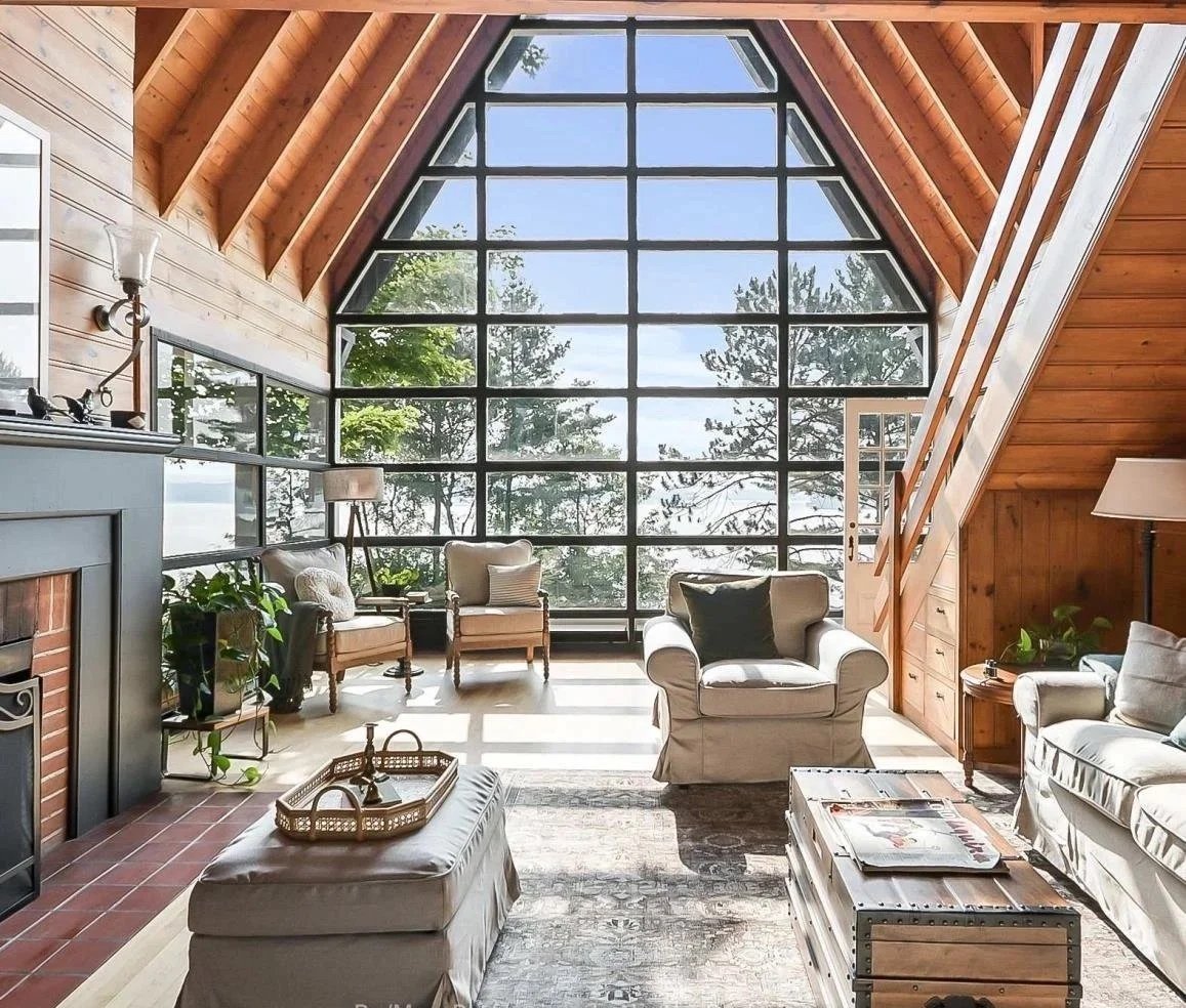Living room with large front-facing windows showing trees and sky, wooden vaulted ceiling, beige furniture, plants, side tables, lamp, fireplace, and a rug.