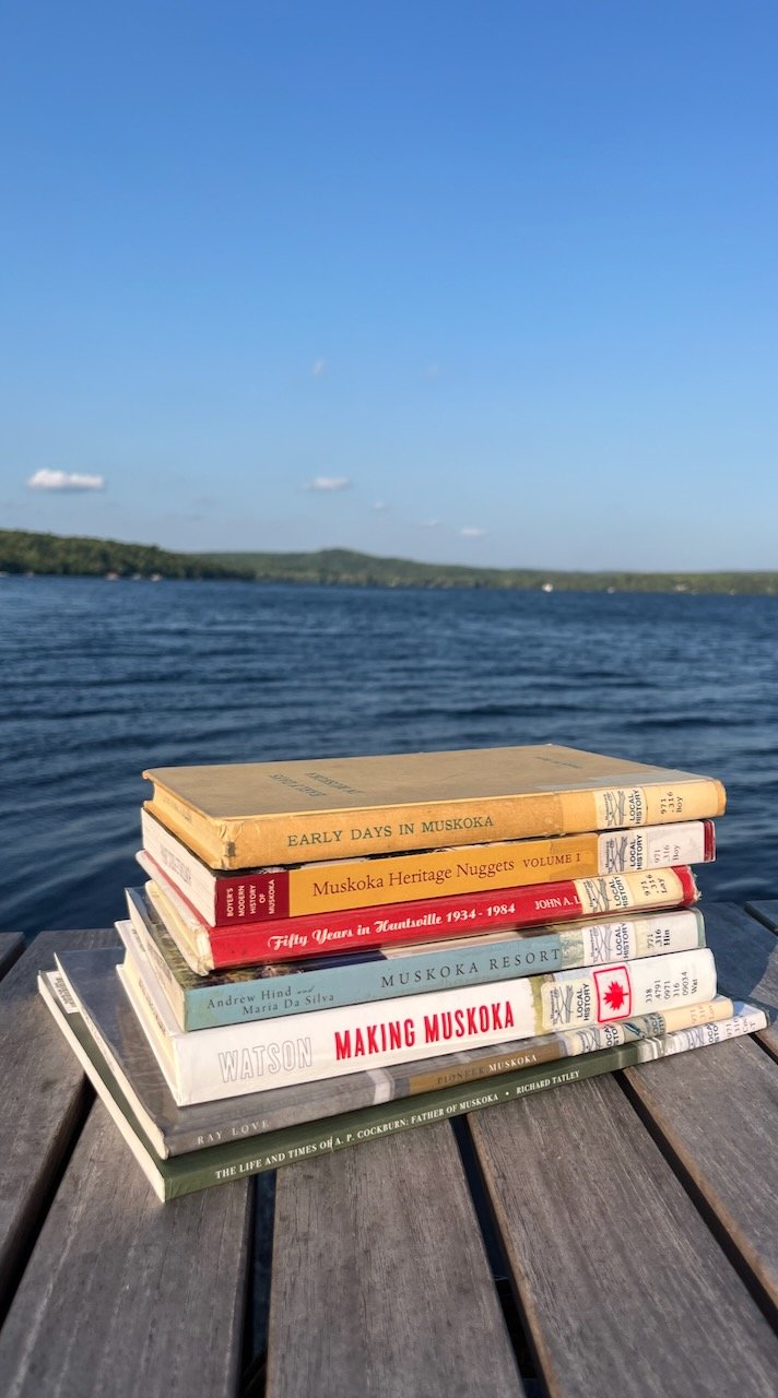 Stack of books about Muskoka, with a lake and green hills in the background.