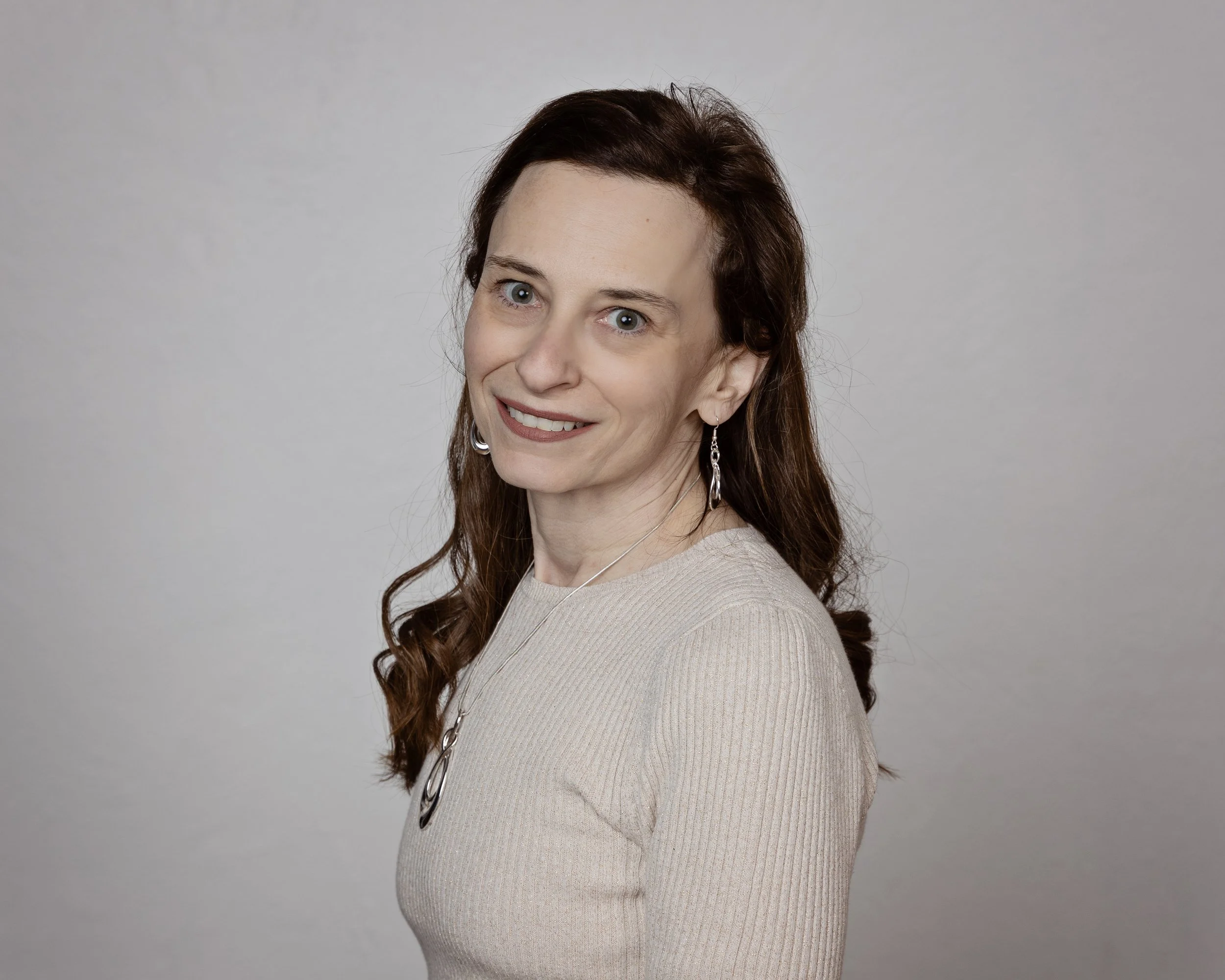 A woman with long curly brown hair wearing a beige sweater, earrings, and a necklace, smiling and looking at the camera against a plain light gray background.