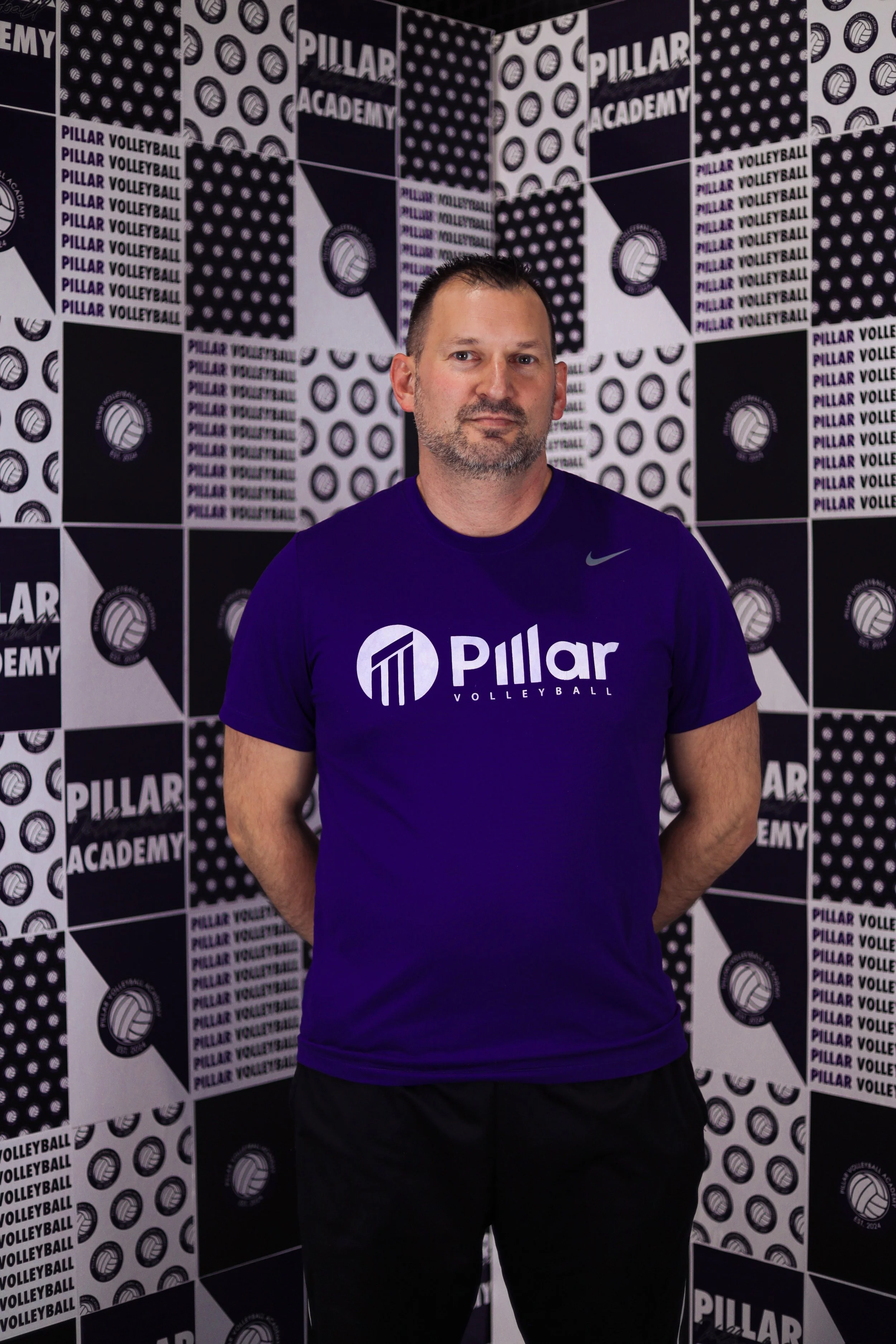 Man standing in front of a wall with black and white Pillar Volleyball branding, wearing a purple Pillar Volleyball T-shirt.