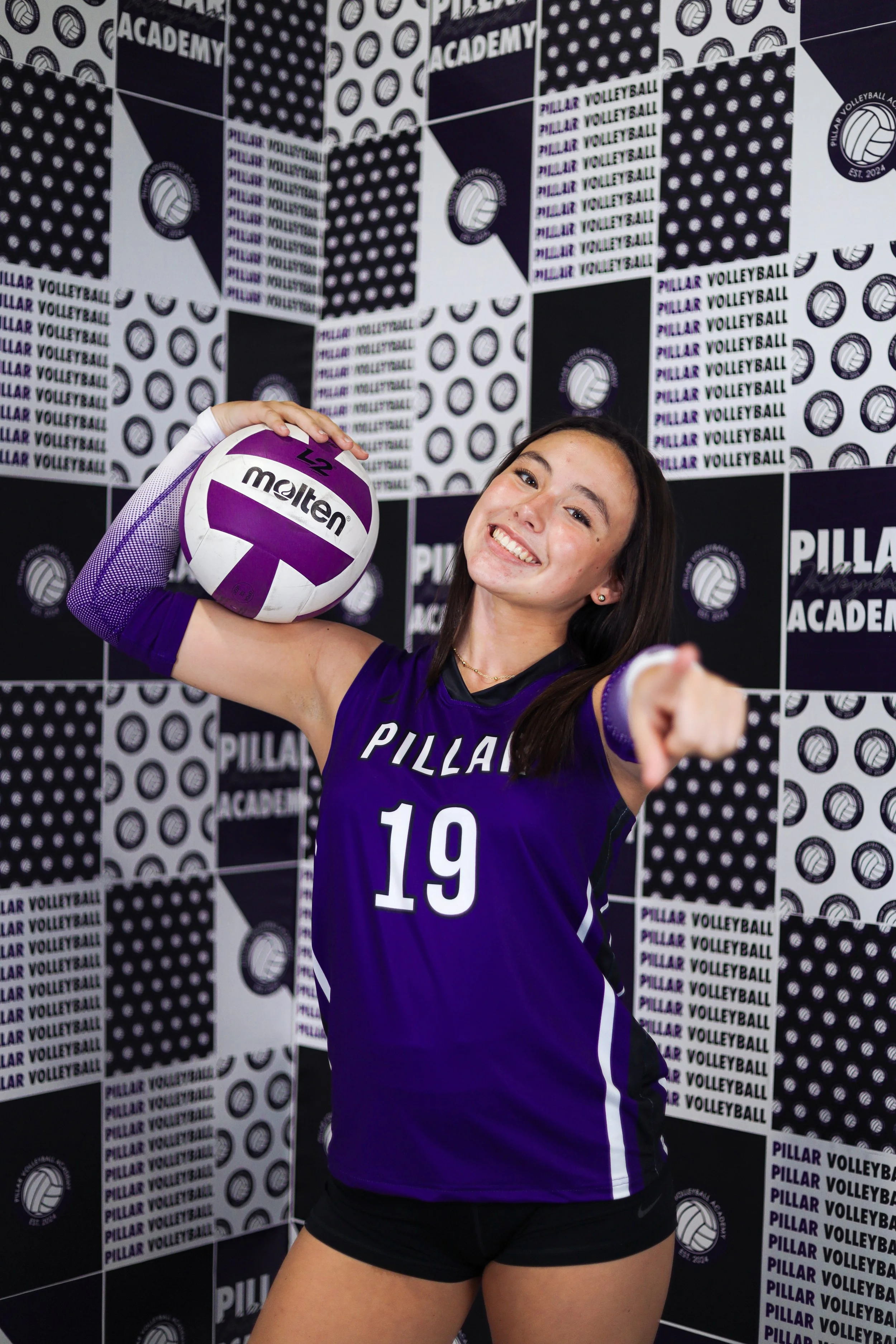 A female volleyball player wearing a purple jersey with the number 19, holding a purple and white Molten volleyball in her right hand and smiling at the camera in front of a black and white patterned backdrop with the words 'Pillar Volleyball Academy.'