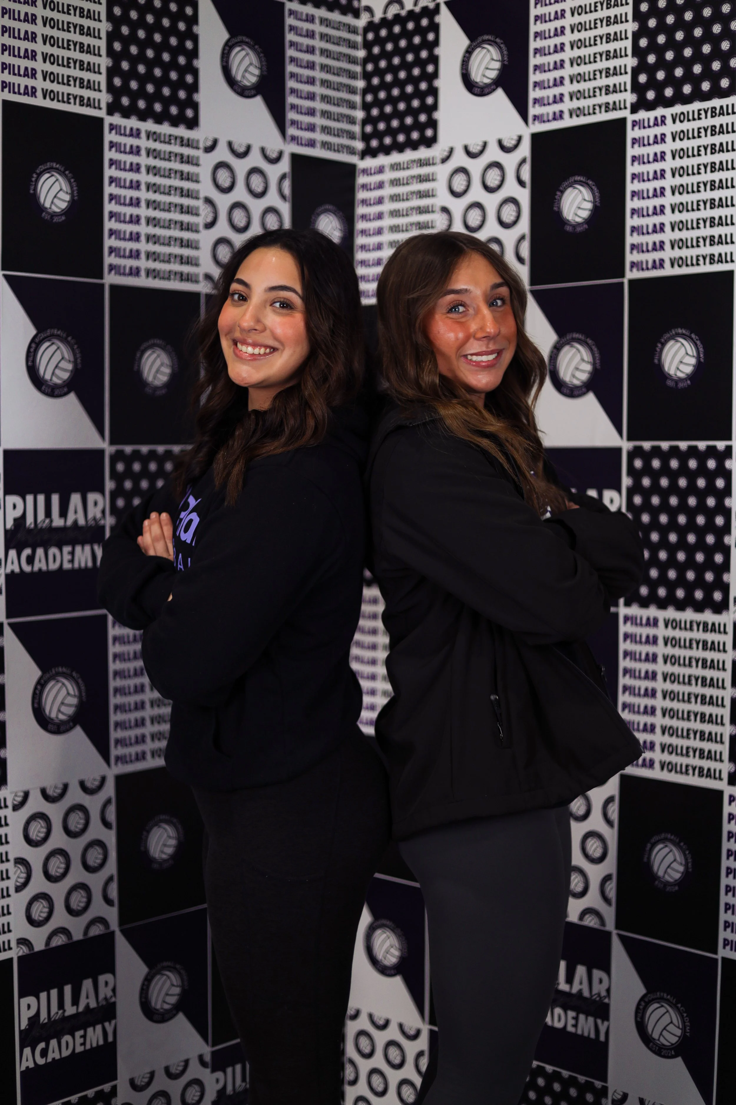 Two women stand back-to-back with crossed arms in front of a black and white volleyball-themed background with text that reads "Pillar Volleyball Academy".