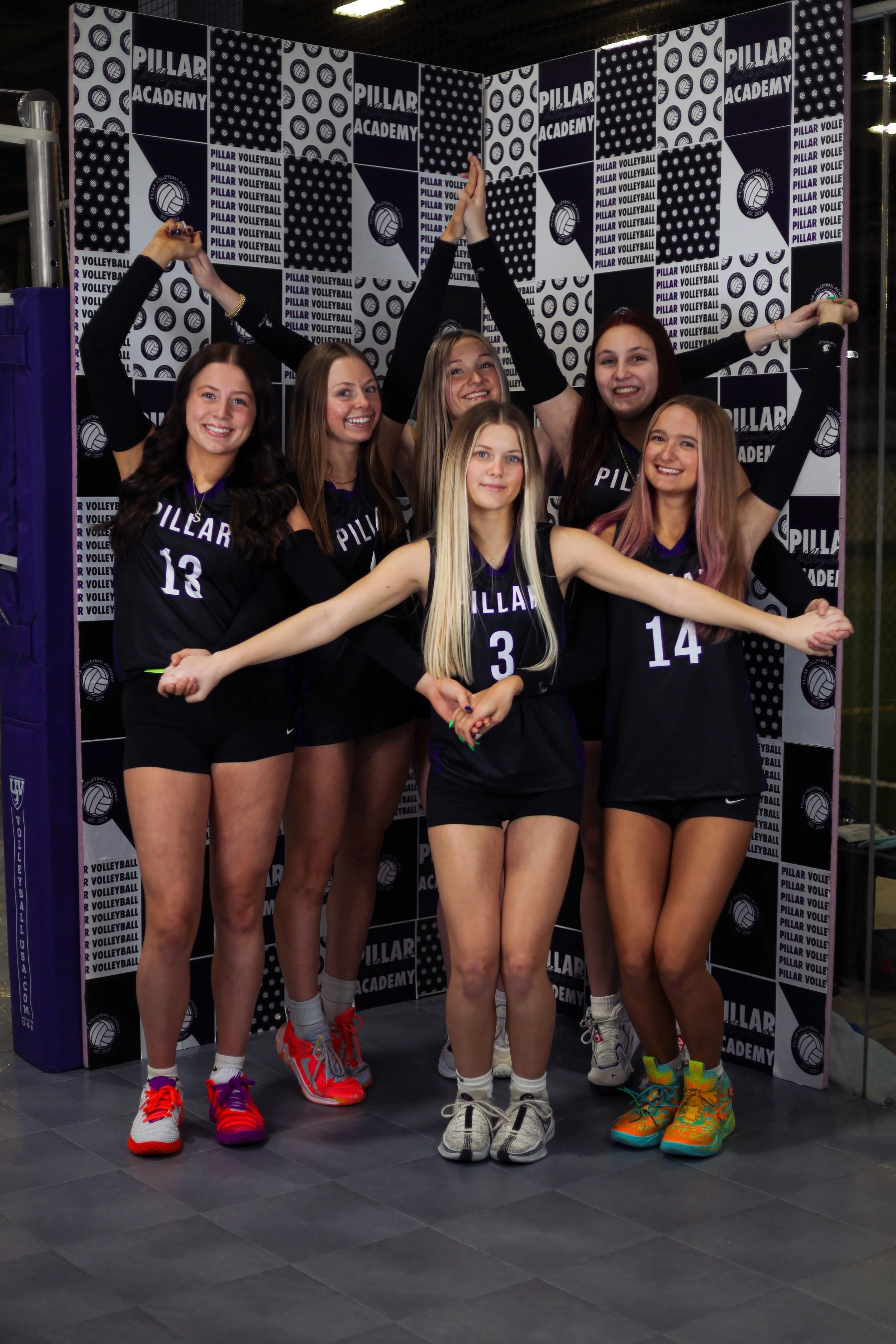 Six female volleyball players in black uniforms with purple accents posing indoors at the Pillar Volleyball Academy, with a black and white pillar-themed backdrop.