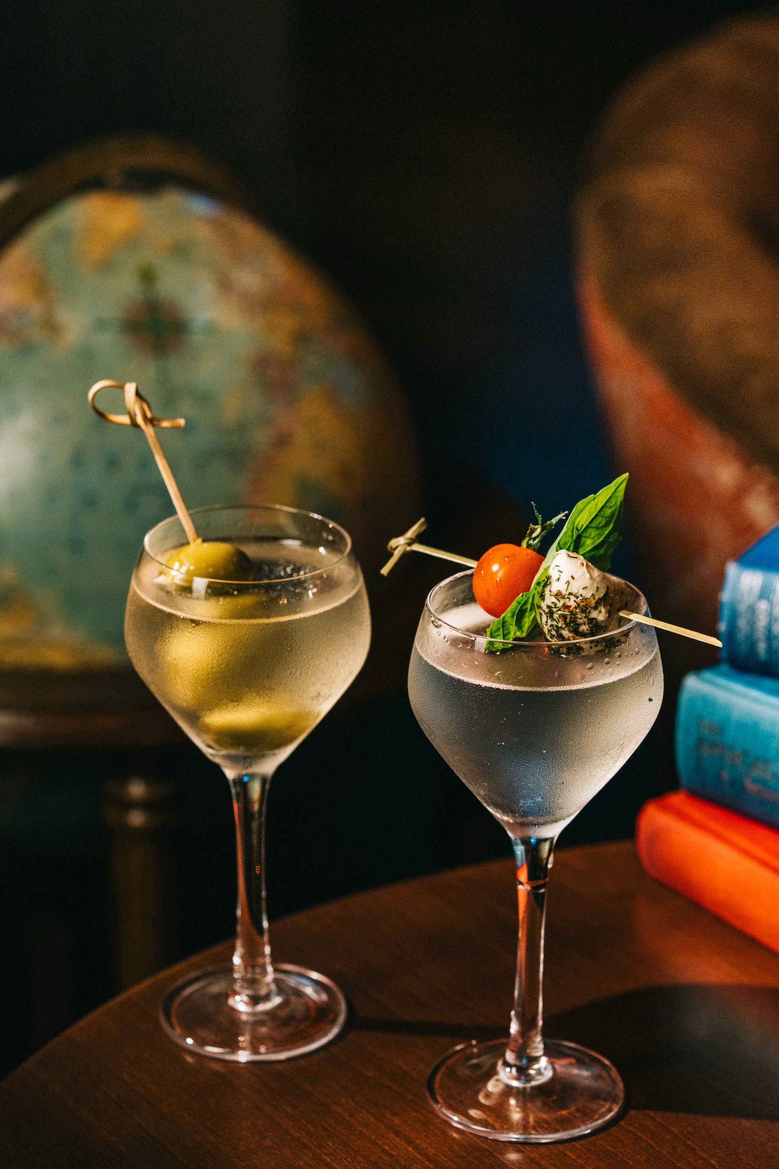 Two cocktails on a wooden table, one garnished with a cherry tomato, basil leaf, and cheese, and the other with an olive, against a backdrop of a world globe and stacked books.