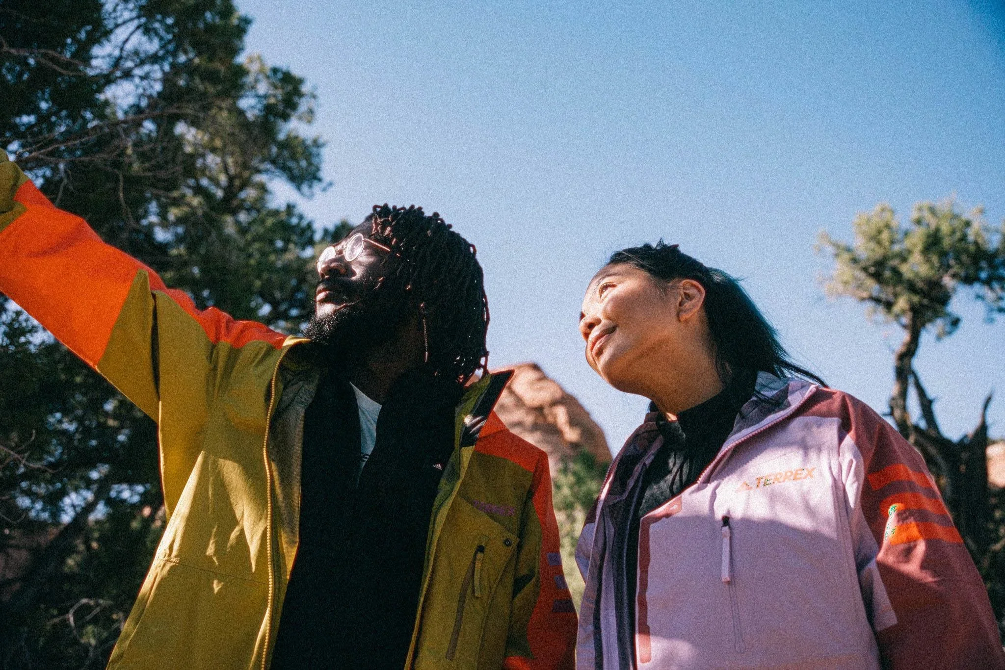 Two friends wearing outdoor jackets standing outdoors under a clear blue sky, enjoying the sunny day.