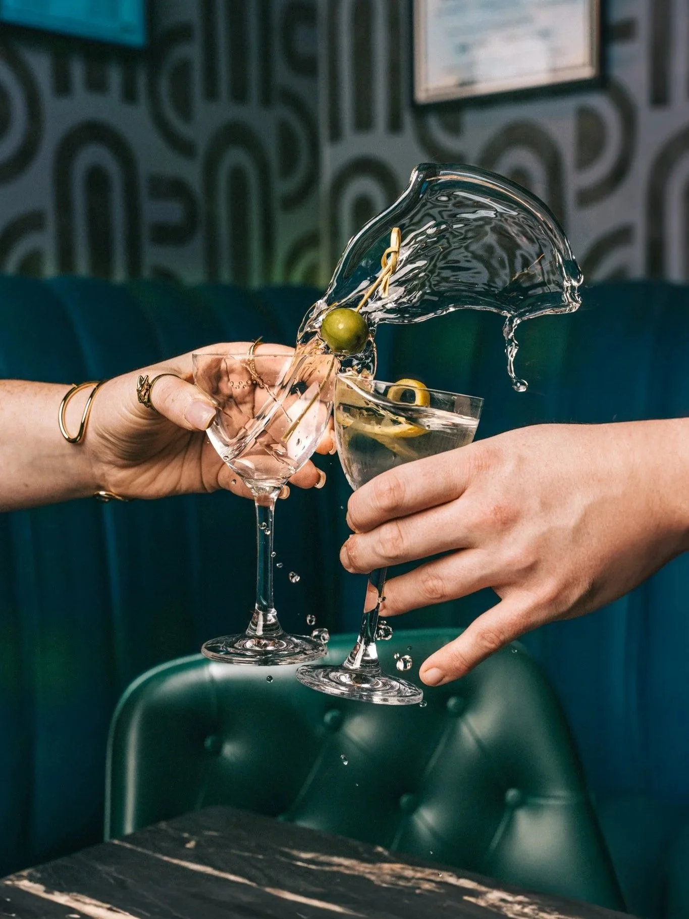 Two people clinking martini glasses with green olives while a splash of liquid flies through the air.