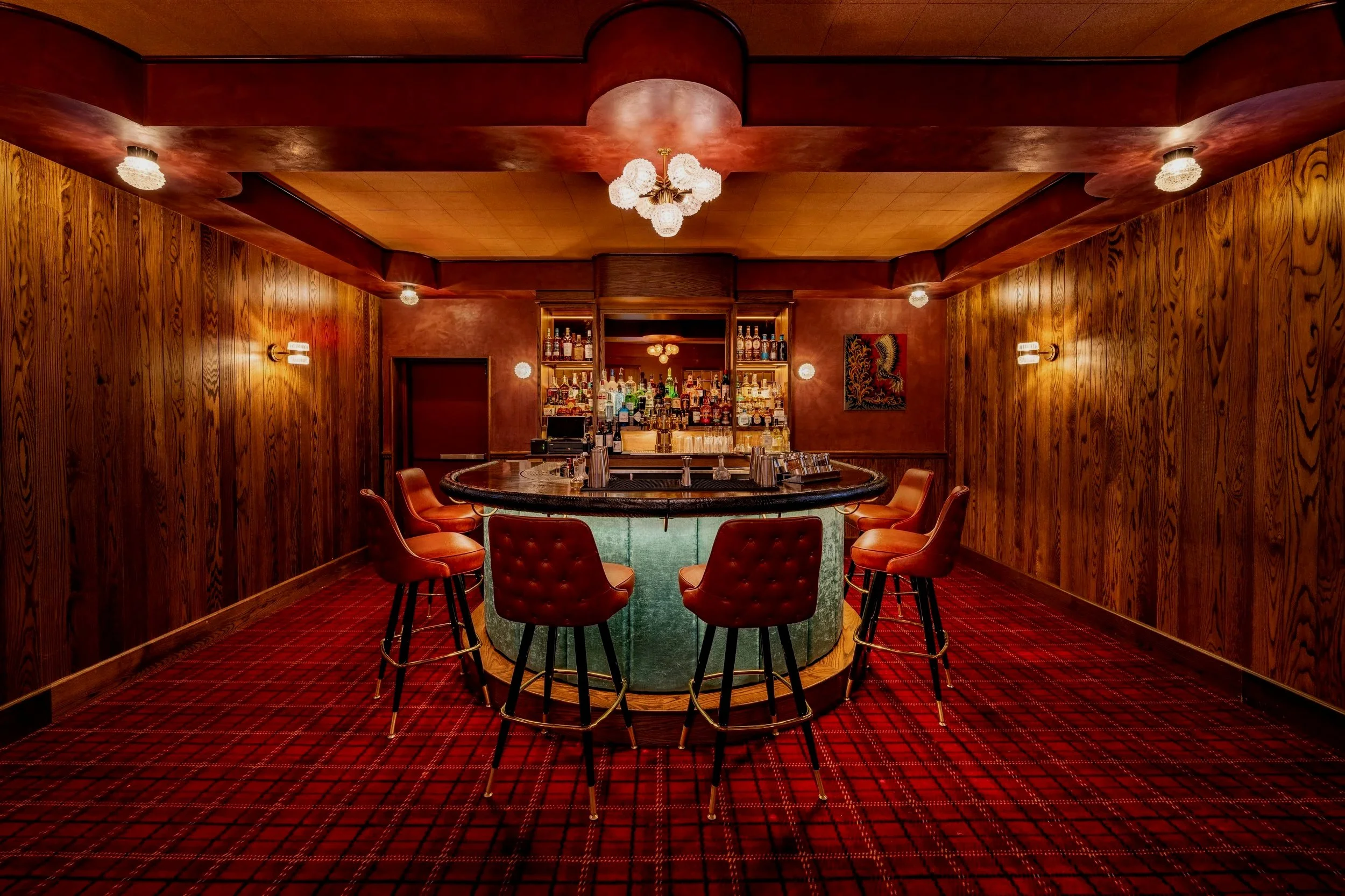 A retro-style bar with a curved green marble bar counter, surrounded by red leather bar stools, wooden paneled walls, a patterned red carpet, and a well-stocked bar with various bottles behind it.