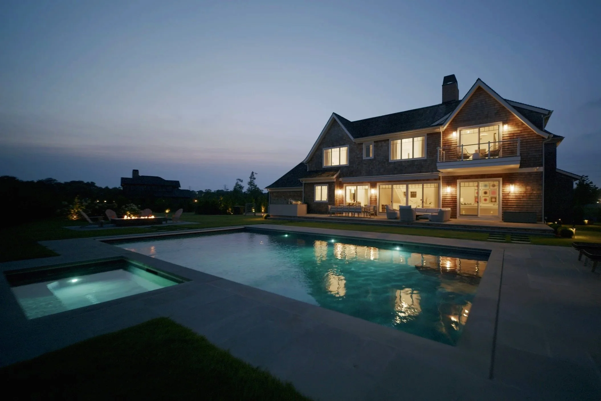 A large house with multiple floors illuminated at dusk, featuring a backyard with a swimming pool and spa, outdoor seating, and a firepit area.