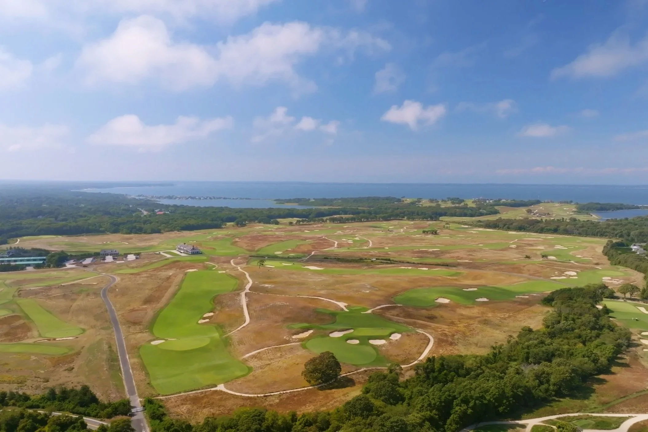 Aerial view of a golf course with lush green fairways and sand traps, surrounded by wooded areas, with a lake and distant shoreline under a partly cloudy sky.