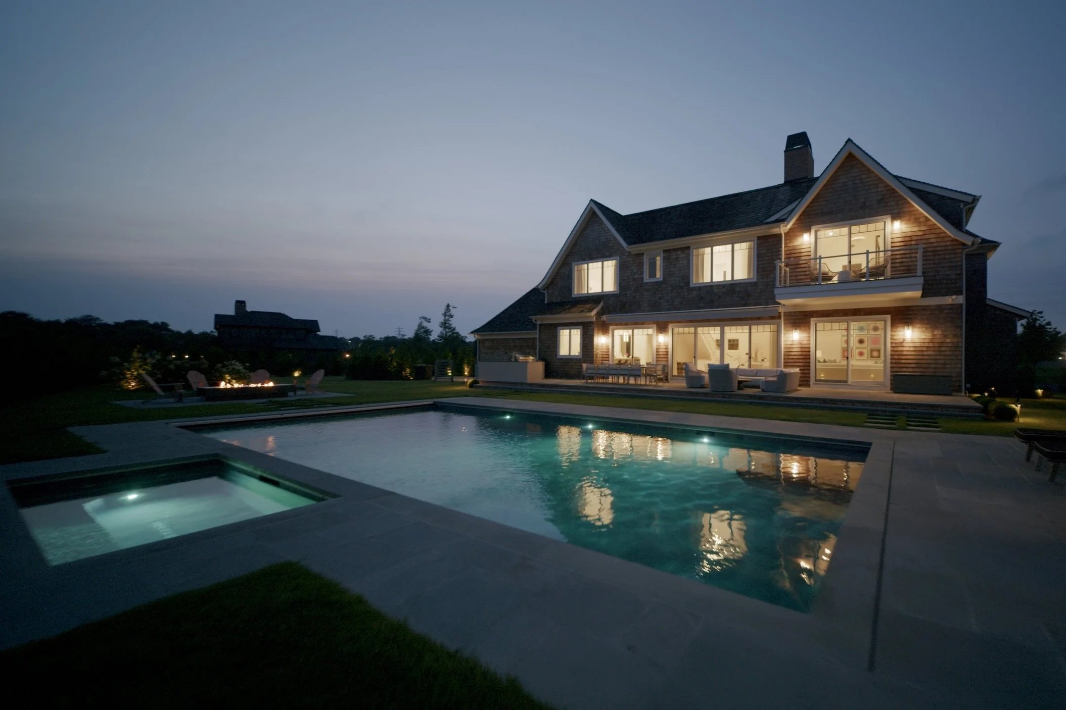 A large house with multiple windows and a balcony, illuminated from inside, is shown at dusk. In the backyard, there is a pool with underwater lights and a hot tub nearby. Outdoor seating areas and a fire pit with chairs are also visible.