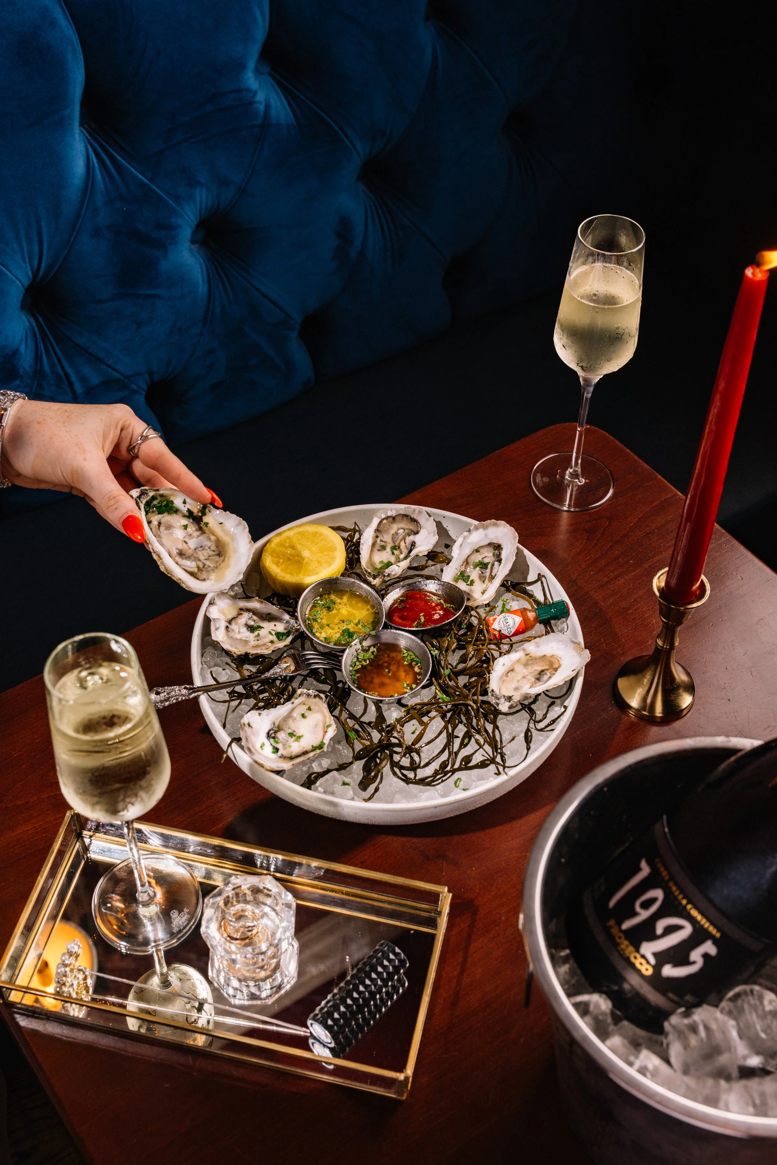 A table with a platter of oysters on the half shell, lemon, various sauces, and seaweed, surrounded by glasses of champagne, a lit red candle, and a bottle of champagne in an ice bucket.