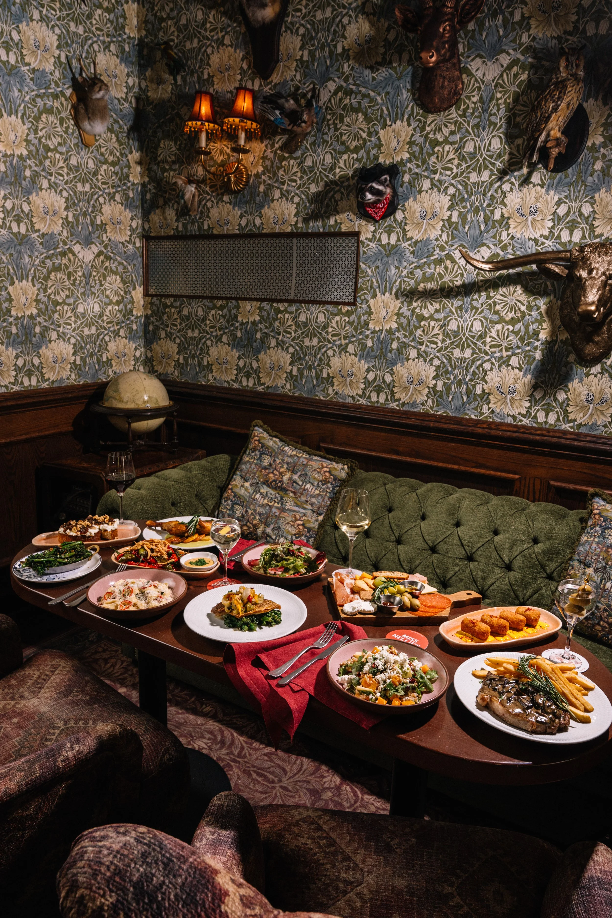 A dining room with a table filled with various dishes, glasses of wine, and cutlery. The background features a green velvet sofa with patterned pillows, a patterned wallpaper with mounted animal heads, and a wall sconce with two lamps.