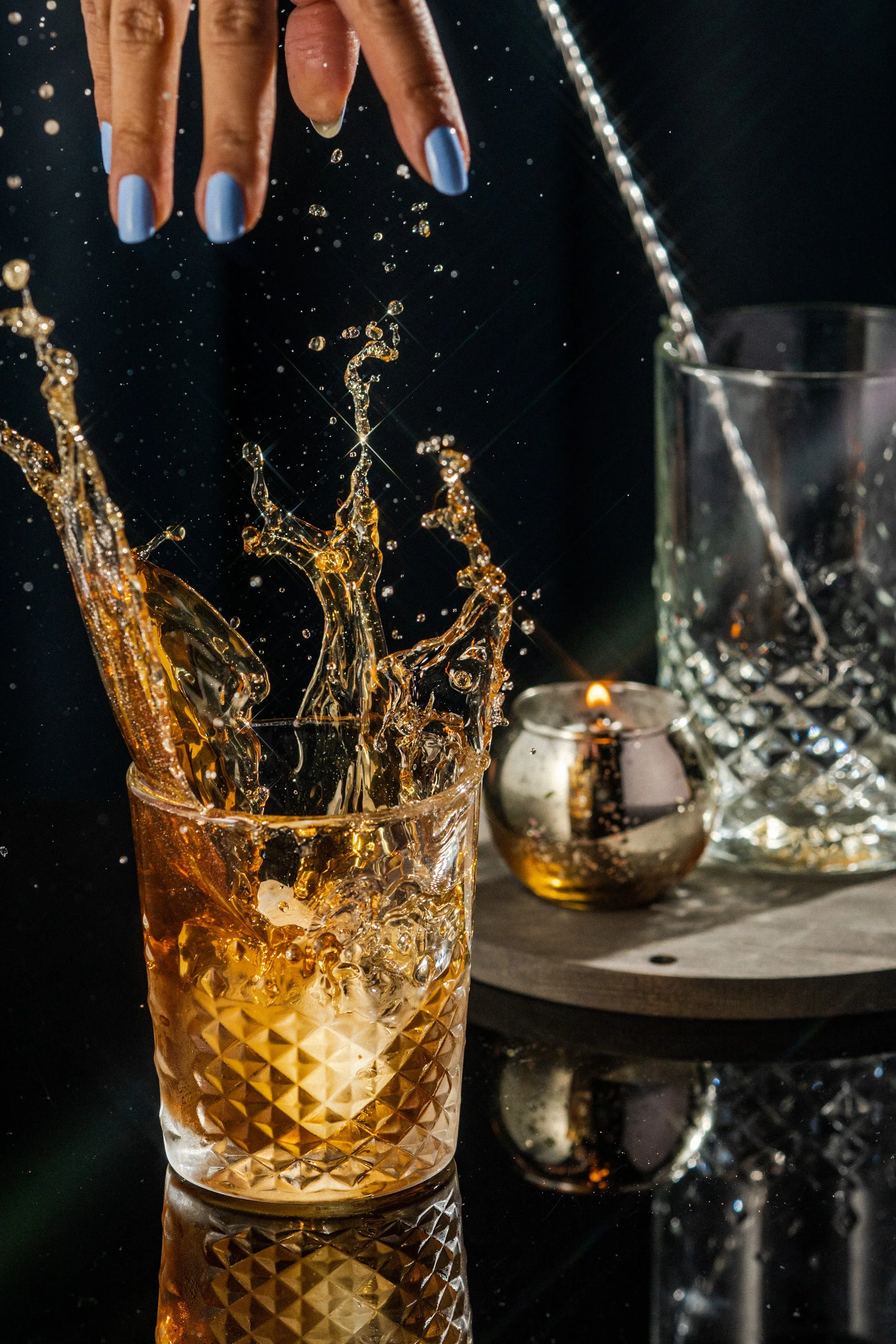 A hand with blue painted nails splashes a drink into a glass, causing water and ice to splash upward. There is a tall glass, a smaller silver container, and a crystal decanter in the background.