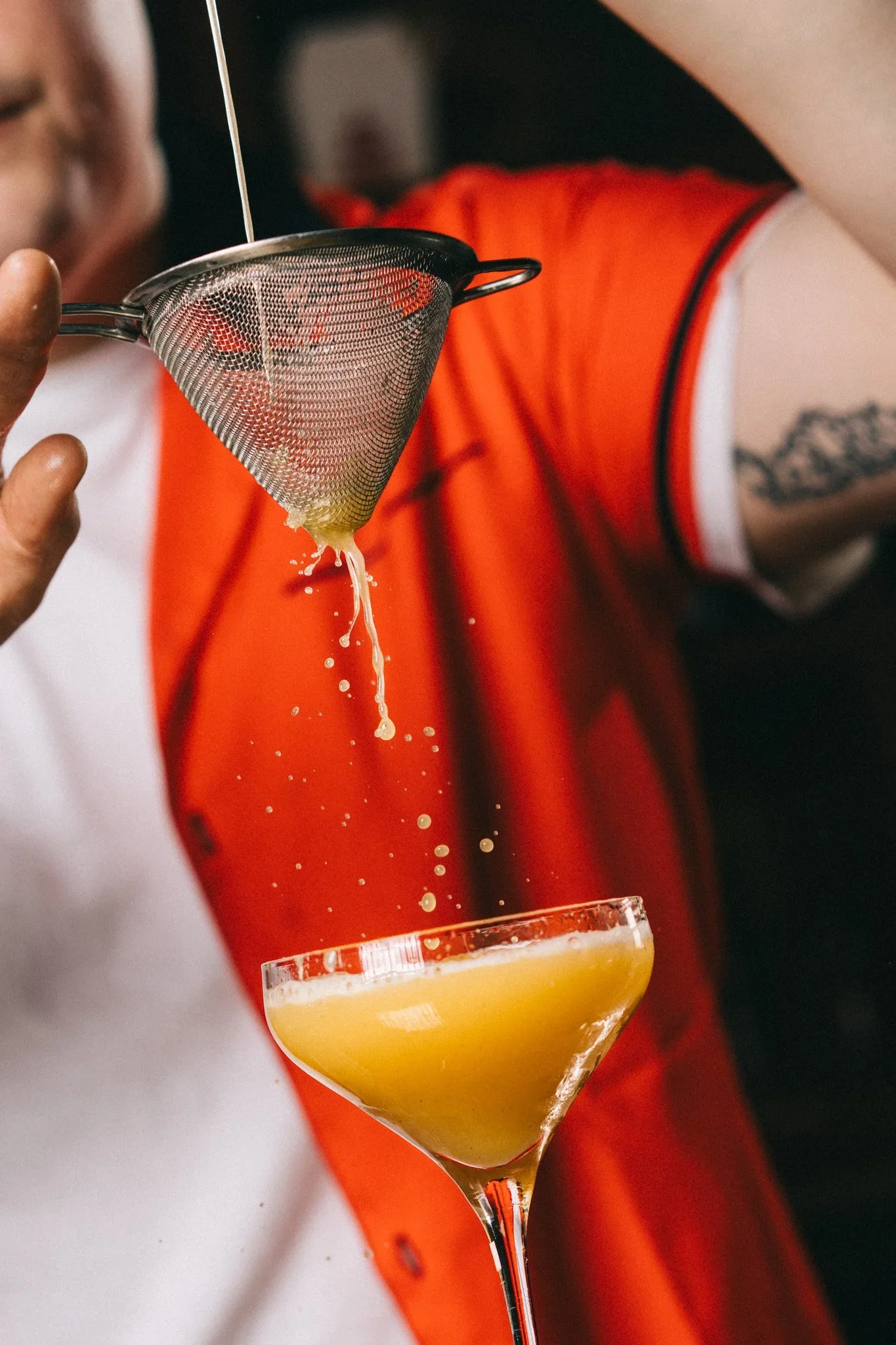 Person pouring a yellow cocktail through a strainer into a cocktail glass.