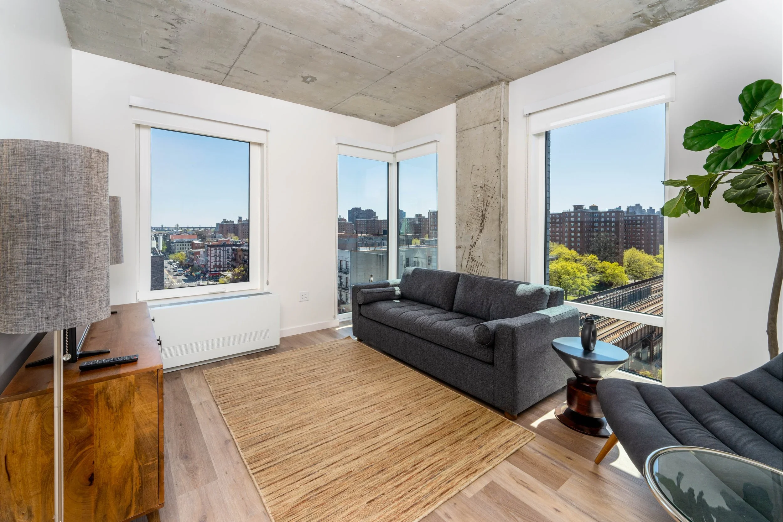 Living room with large windows showing cityscape, modern furniture including a dark gray sofa and a matching chair, a wooden side table with a vase and a small round table, a textured rug, a potted plant, and a concrete ceiling.