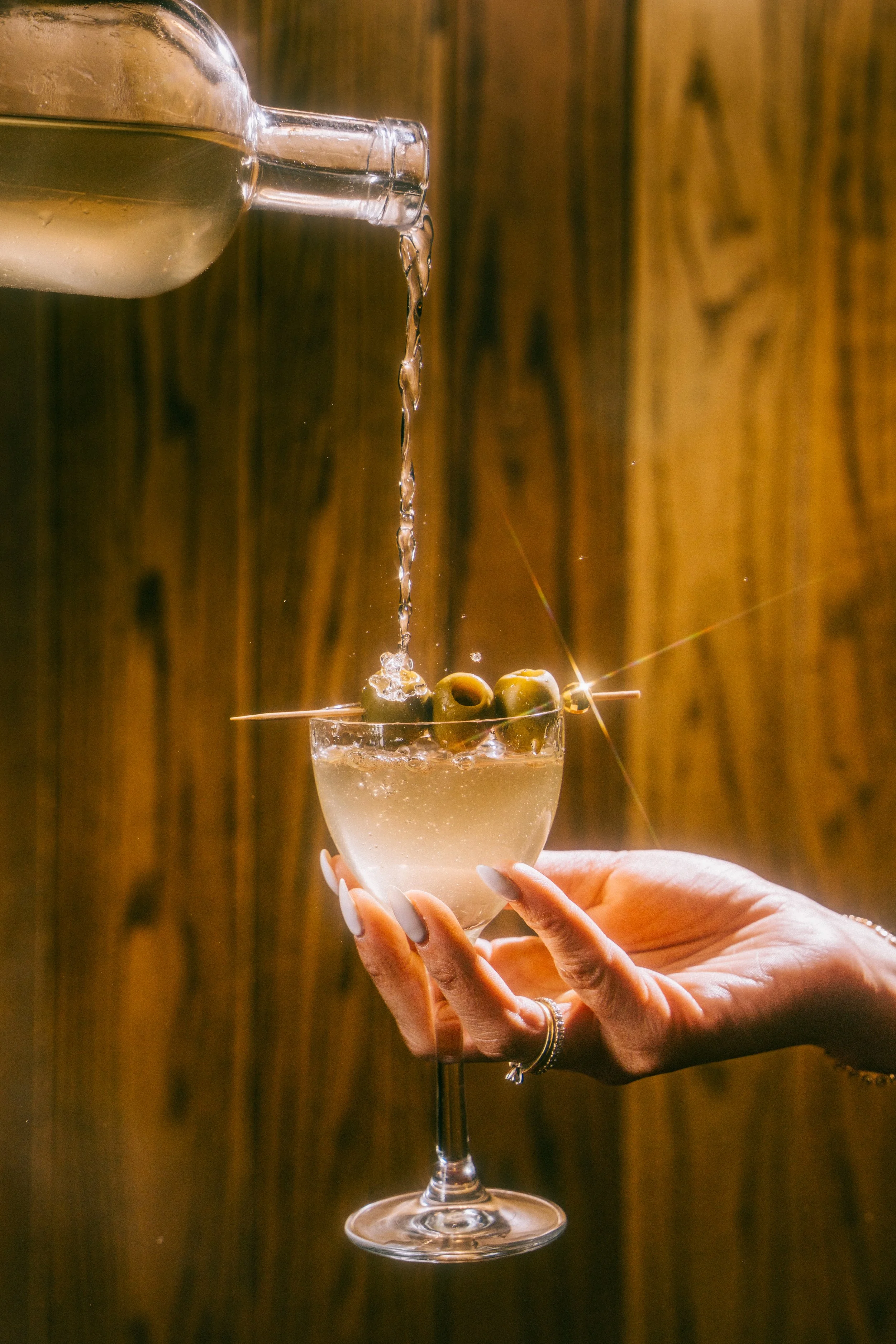 A hand holding a stemmed glass with drinks, while water is being poured from a bottle into the glass with green olives on a skewer, against a wooden background.