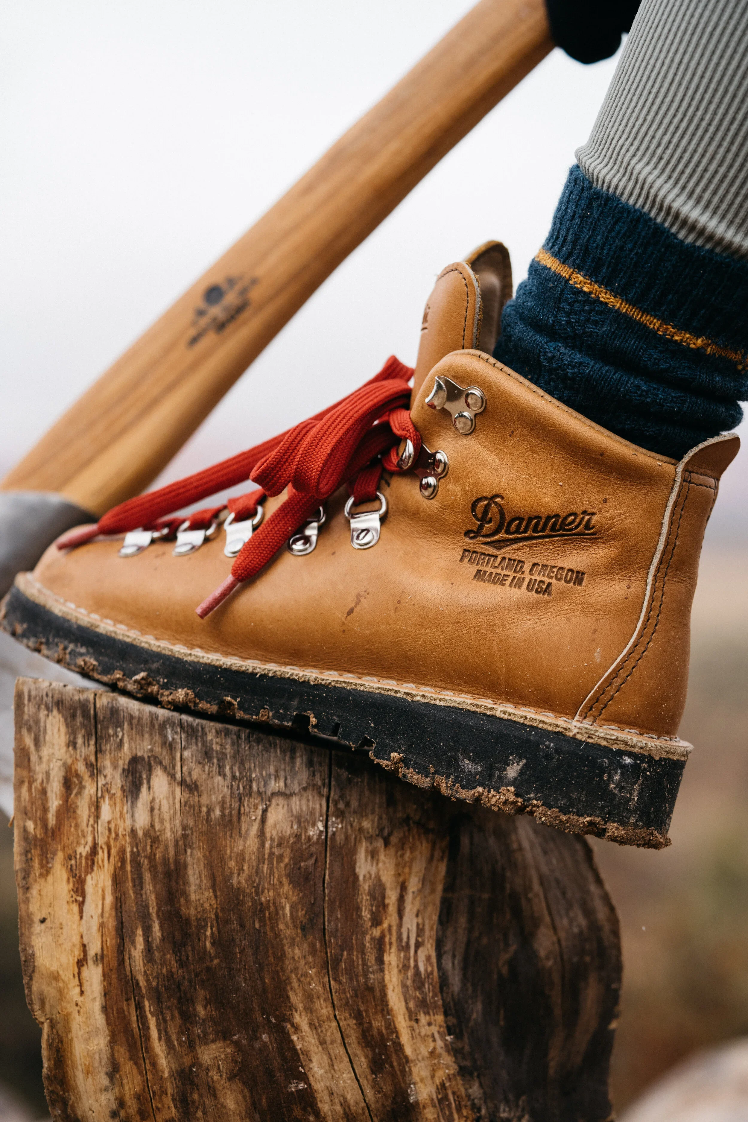 Close-up of a tan leather Danner hiking boot with red laces, worn on a person's foot. The boot is resting on a weathered wooden stump, with an axe handle leaning against the person's leg. The person is wearing jeans and a striped sock.