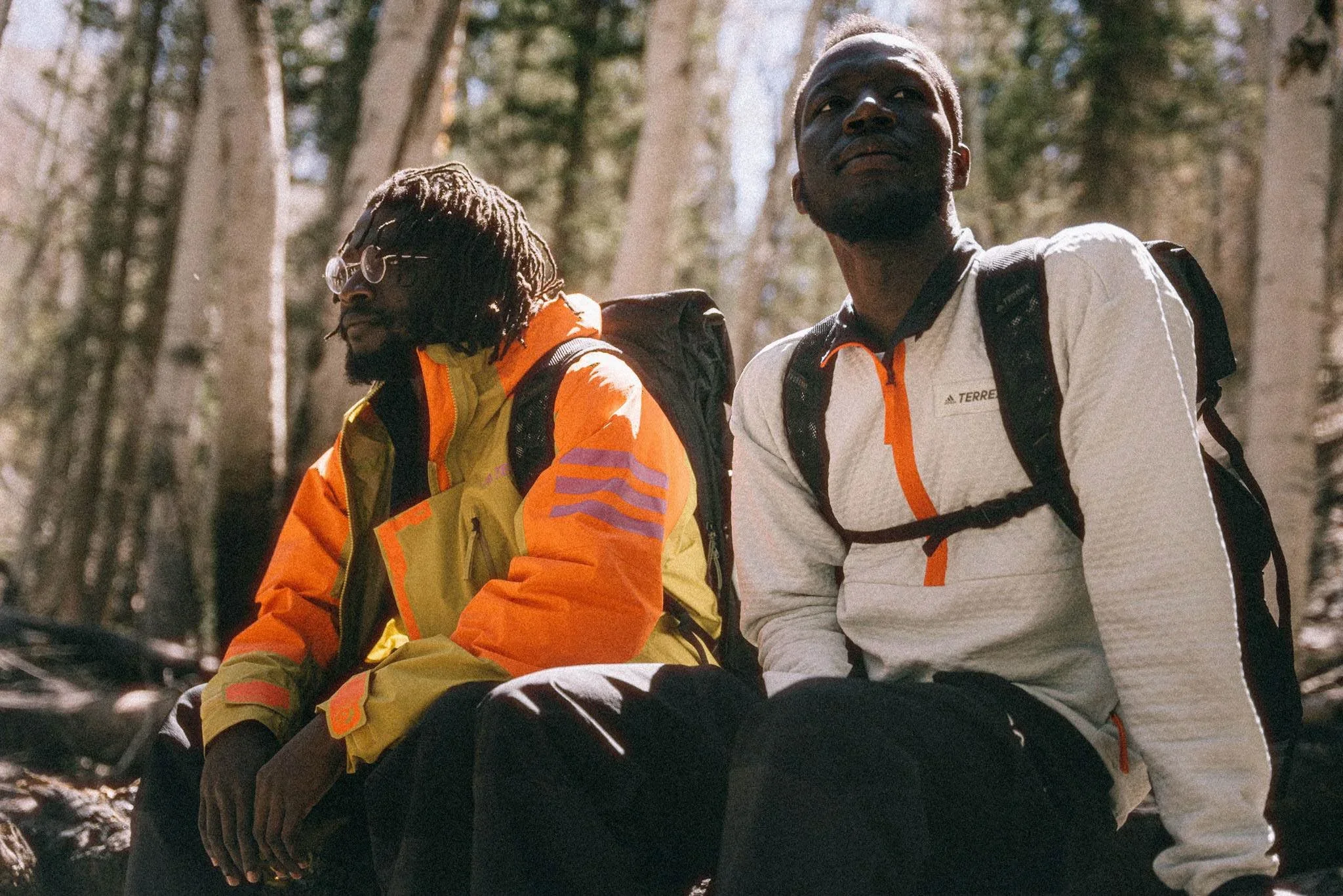 Two men sitting on a log in a forest, wearing backpacks and outdoor clothing, with trees in the background.