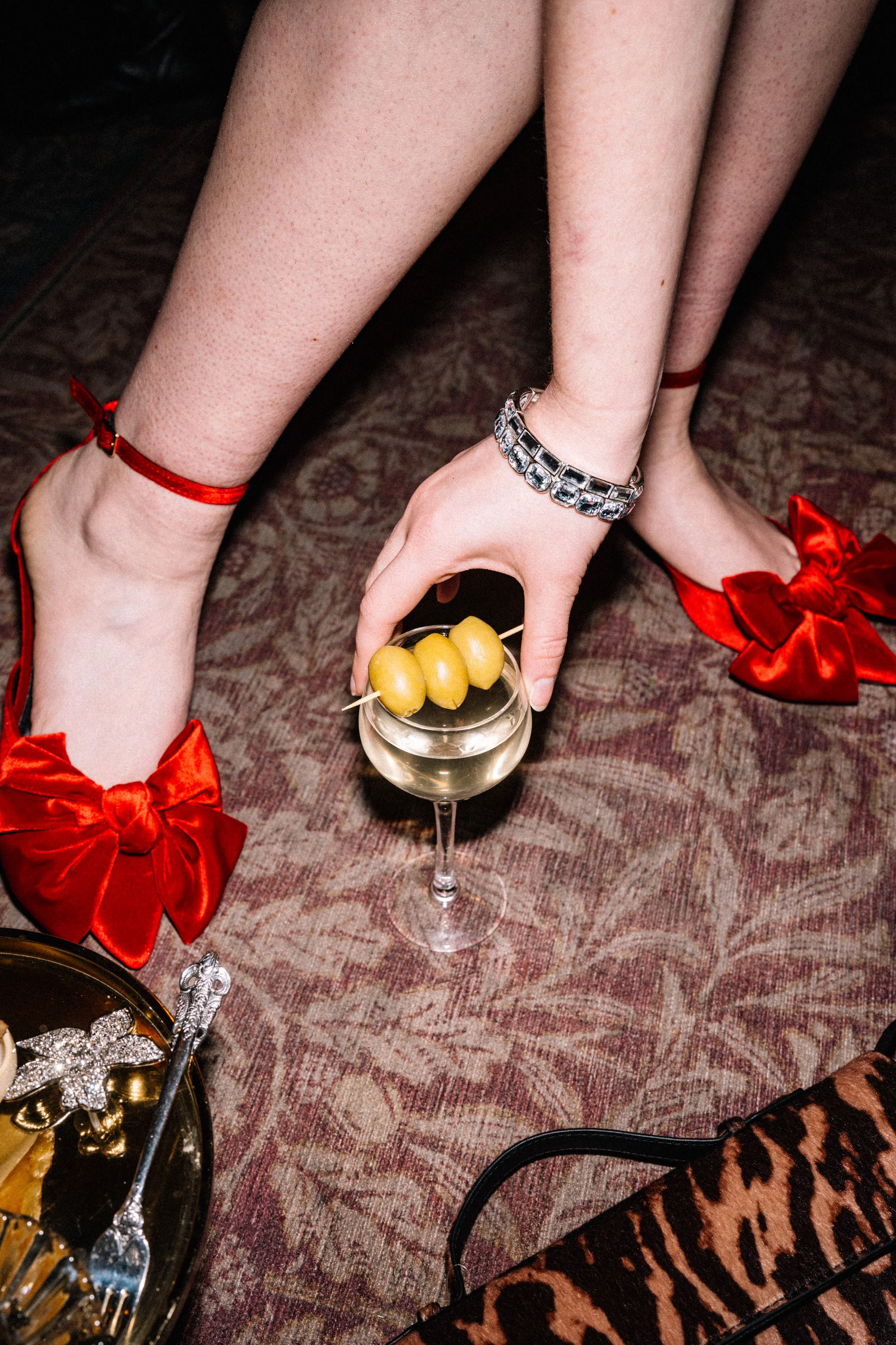 A person is adjusting a champagne glass with green olives on a skewer. The person is wearing red satin shoes with large bows, fishnet stockings, a bracelet, and red anklets. The setting appears to be a party or celebration.