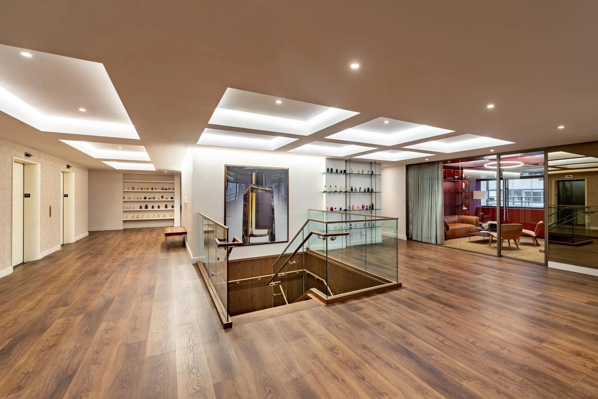 Modern interior of a hotel lobby or lounge area with wooden flooring, ceiling lights, elevator doors on the left, glass railing around stairwell, and a seating area with a sofa and chairs visible through glass walls on the right.