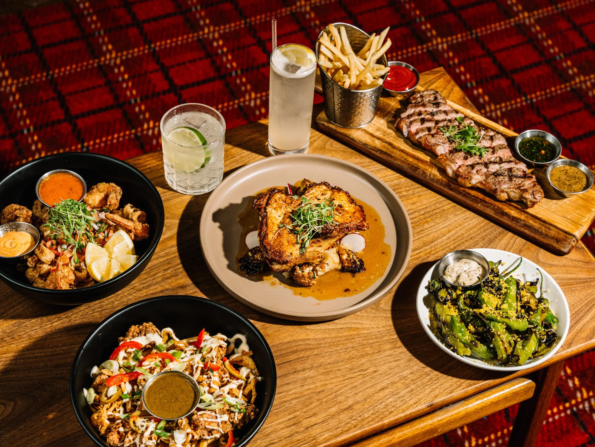 A variety of dishes on a wooden table including fried chicken, grilled chicken, steak with fries, a vegetable salad, and a mixed salad, along with drinks and sauces.