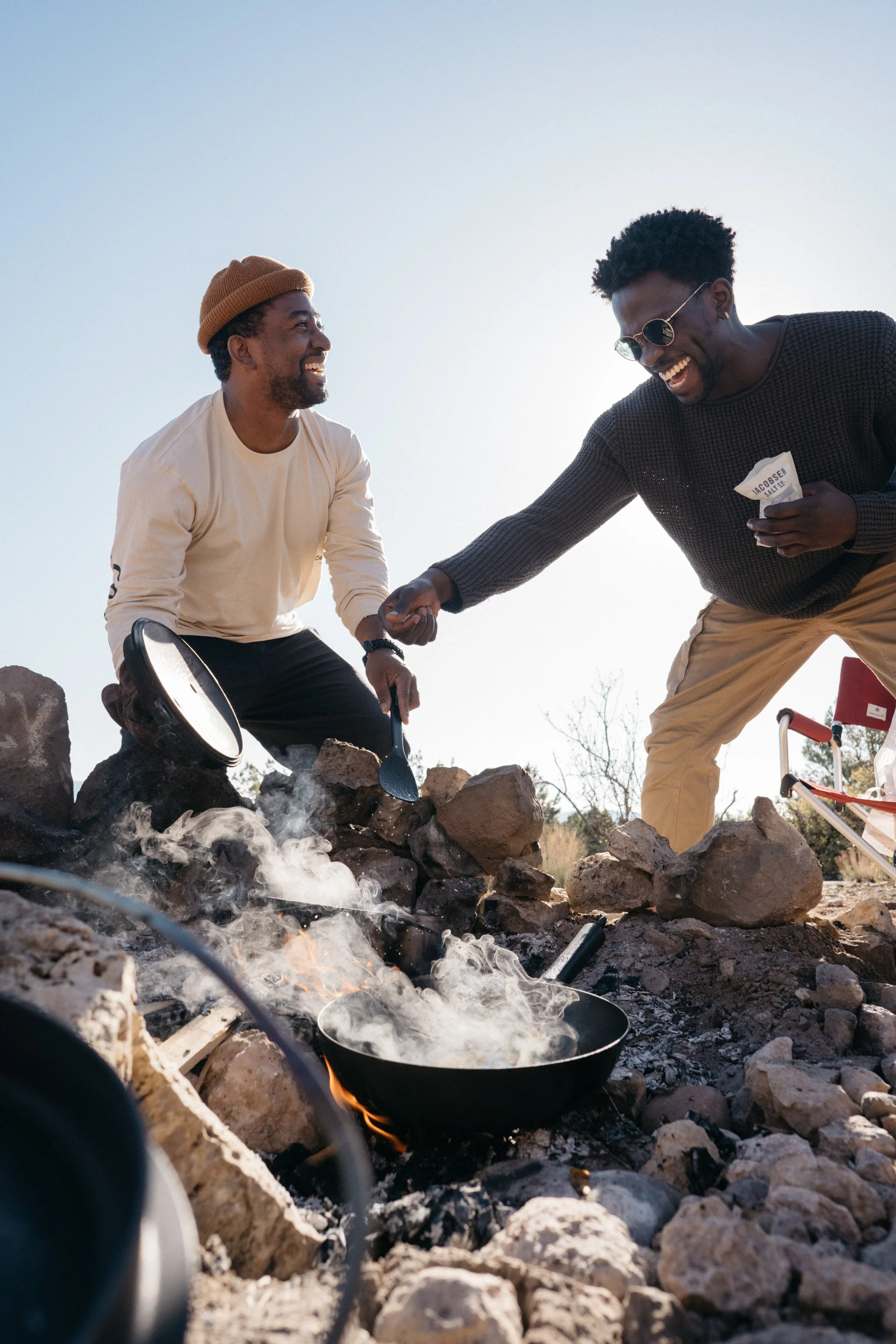 Two men camping outdoors near a campfire, cooking in a frying pan, smiling and holding hands.