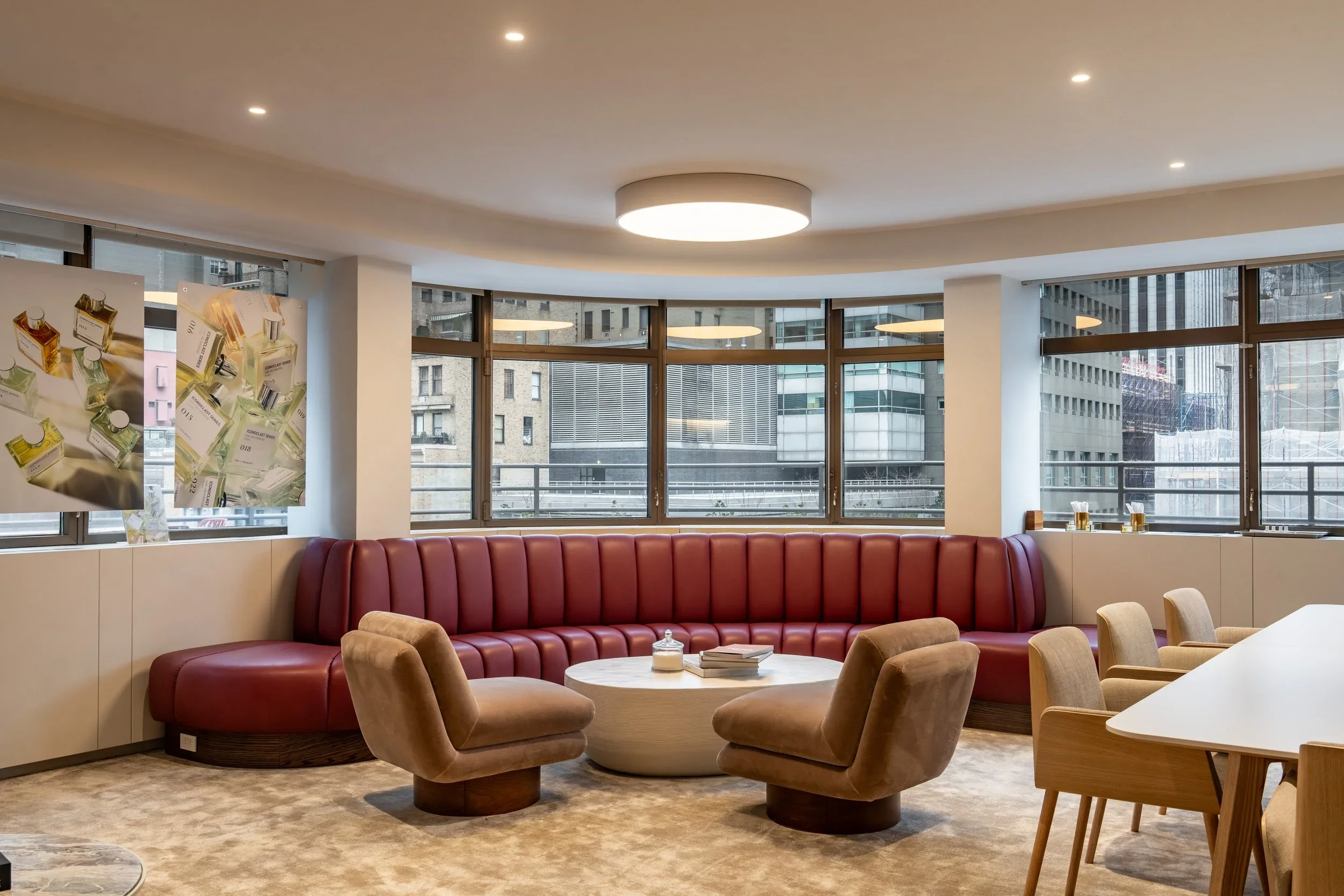 Modern lounge with red curved sofa, beige armchairs, round coffee table with books and decorative items, large windows with cityscape view, abstract artwork on the wall, and ceiling lights.