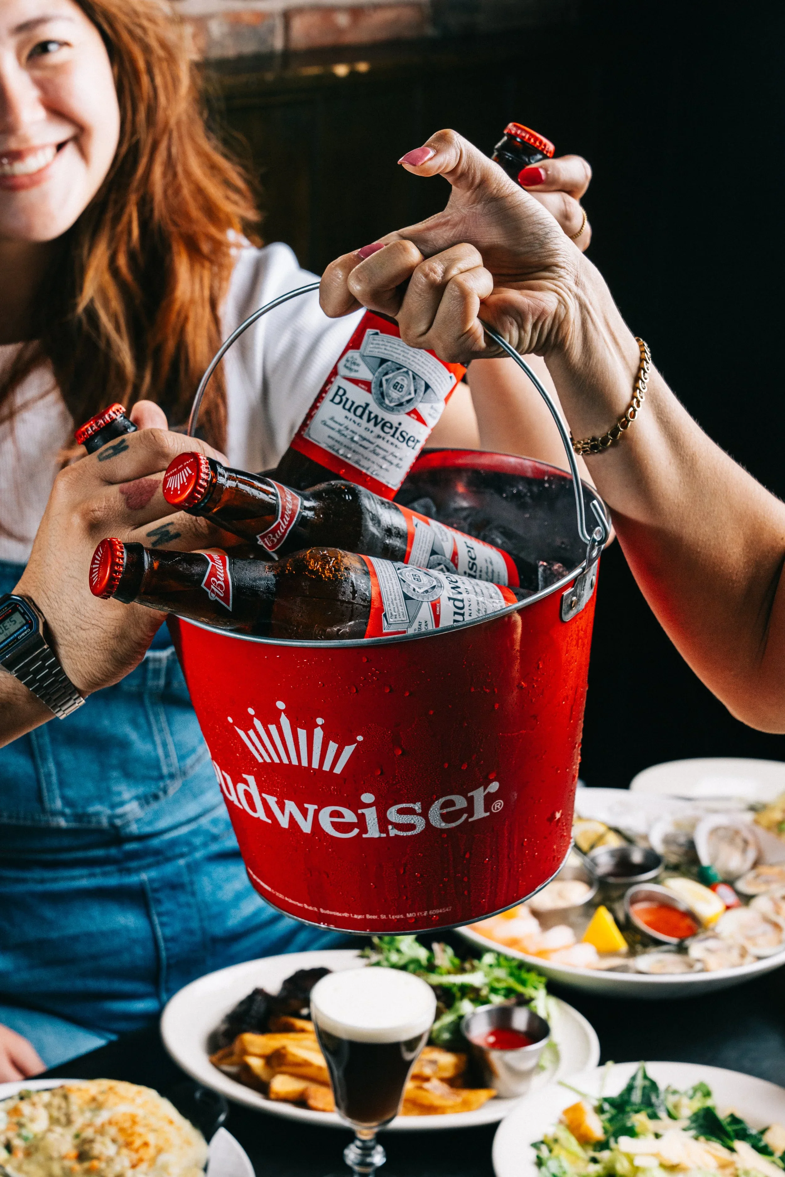 People sharing a bucket of Budweiser beer outdoors with various dishes on the table.